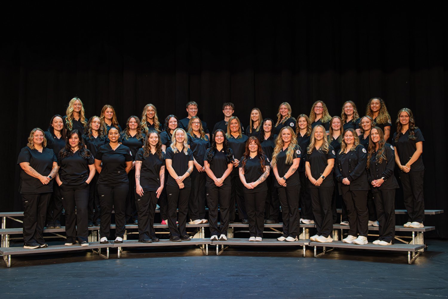 A large group photo of nursing students from the 2026 Associate Degree in Nursing (ADN) program standing on tiered risers on a stage with a black curtain backdrop. All students are wearing matching dark navy or black medical scrubs.  The students are arranged in three rows, from left to right:  Front Row: Tori Trout, Chelsea Bonilla, K-Sha Morgan, Alexis Mendenall, Emily Crews, Kris Kreger, Madison Lane, Karlea Harris, Paige Greenwalt, Lillian Myers, Josie Jackson  Middle Row: Katie Campbell, Alexandria Brown, Kelsey Braden-Stroud, Tylar Helbig, Jade Neal, Cortney Lach, Krista Nickell, Leah York, Morgan Heil, Saylor Lappin, Lexie Sala  Back Row: Hallie Woodrow, Jayla Shelton, Brooklyn Knight, David Woodcock, Michael Parks, Karlie Murphy, Kylie Cockrum, Kaitlyn Worley, Elizabeth Eastham, Shianne Dejear