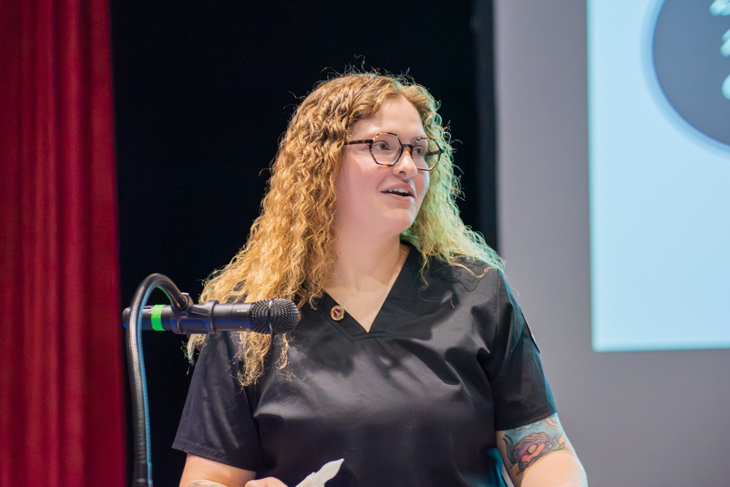 A close-up shot of nursing student Kaitlyn Worley standing at a podium with a microphone on a stage. She has long, curly blonde hair, wears tortoiseshell glasses, and is dressed in black medical scrubs with a nursing pin visible near her collar. A red stage curtain is on the left, and a projection screen is in the background.