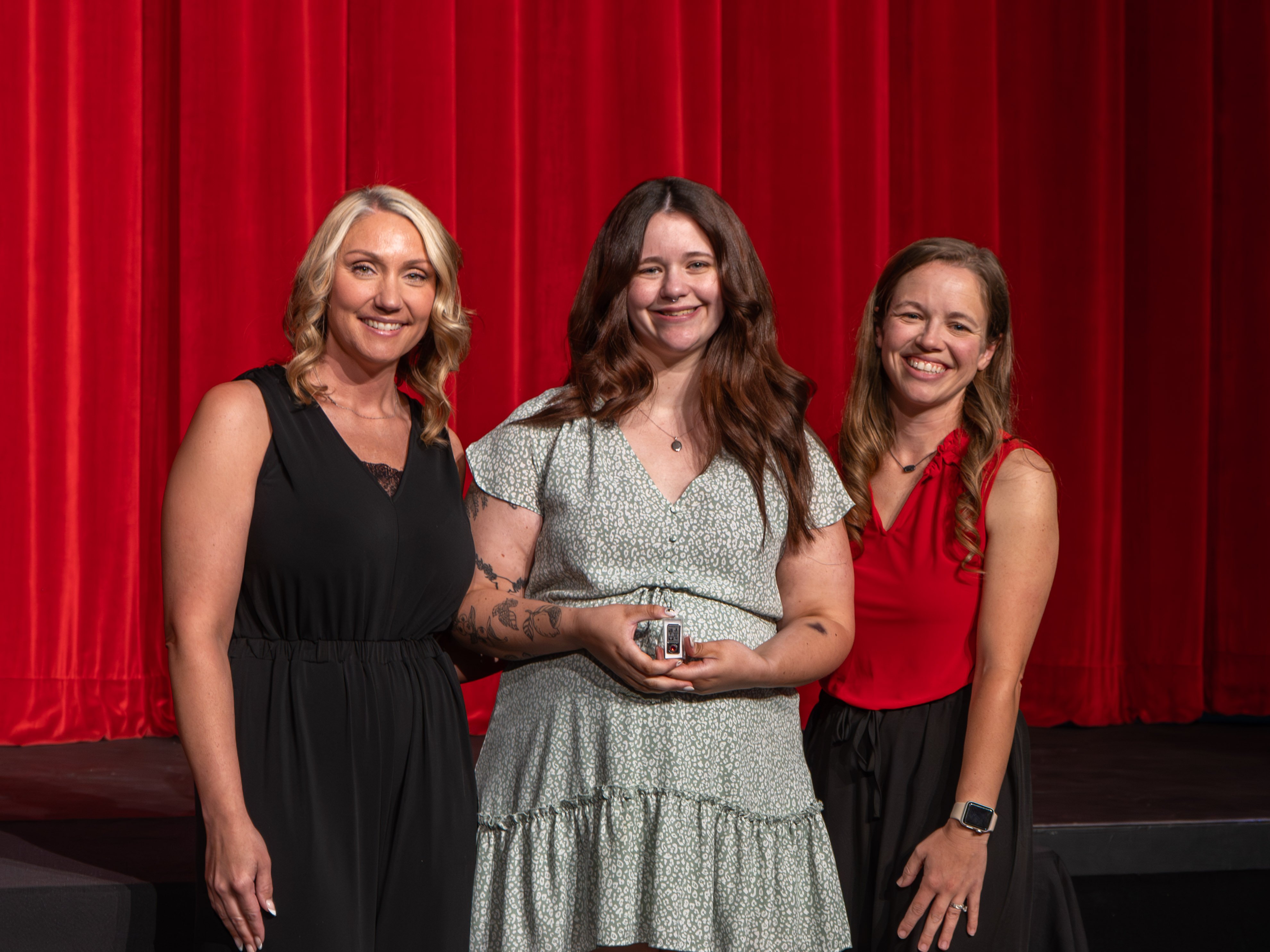 A close-up shot of graduate Allyson Tate, a young woman with long dark hair and botanical arm tattoos wearing a light green floral dress. A male faculty member wearing a grey polo shirt stands to her left, attaching a program pin to her shoulder strap against a red stage curtain background.
