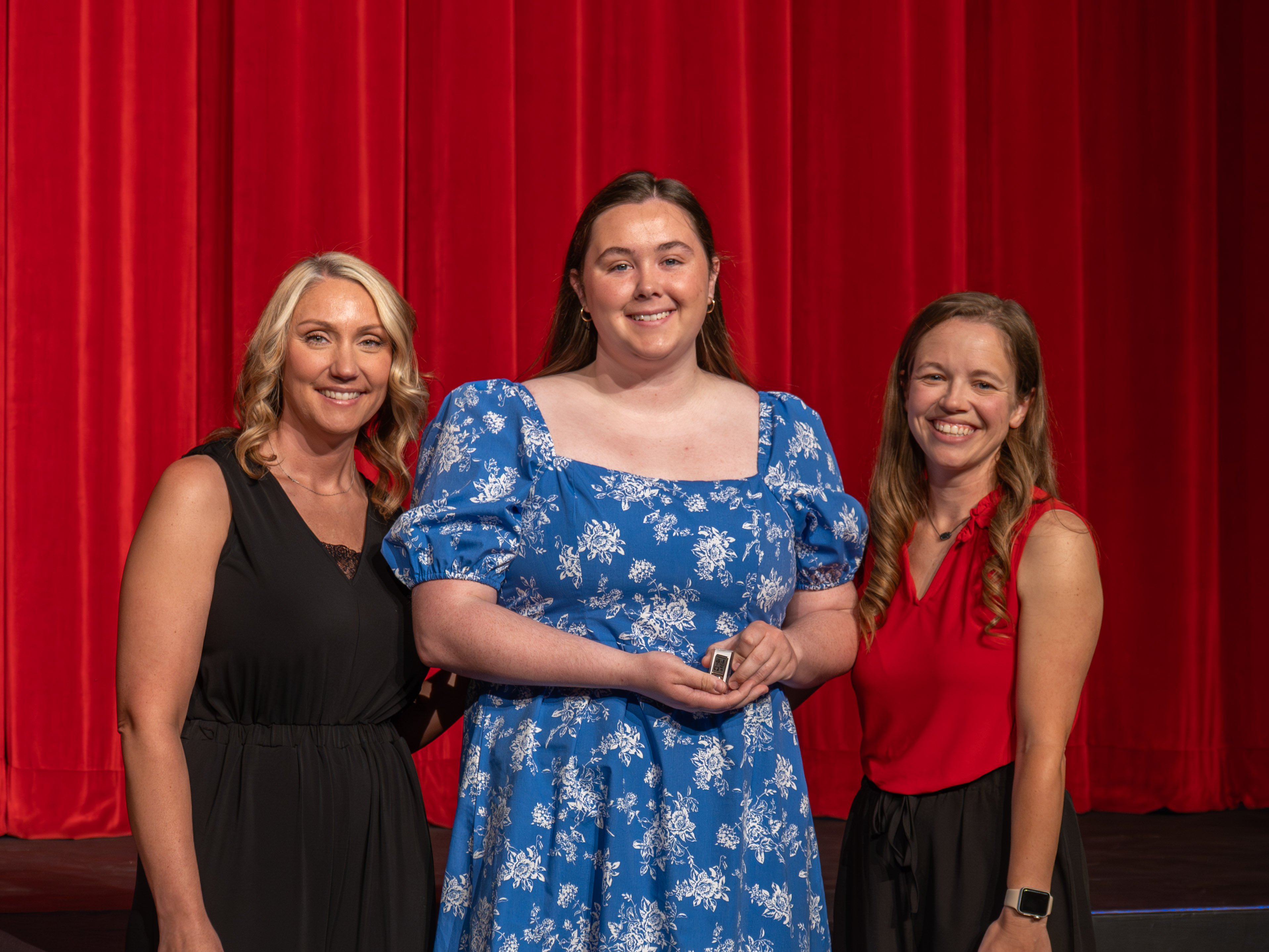 A close-up shot of graduate Caralin Numi, a smiling young woman with straight brown hair wearing a bright blue dress with a white floral pattern. A male faculty member wearing a grey polo shirt stands beside her, placing a program pin on her shoulder strap in front of a red stage curtain.