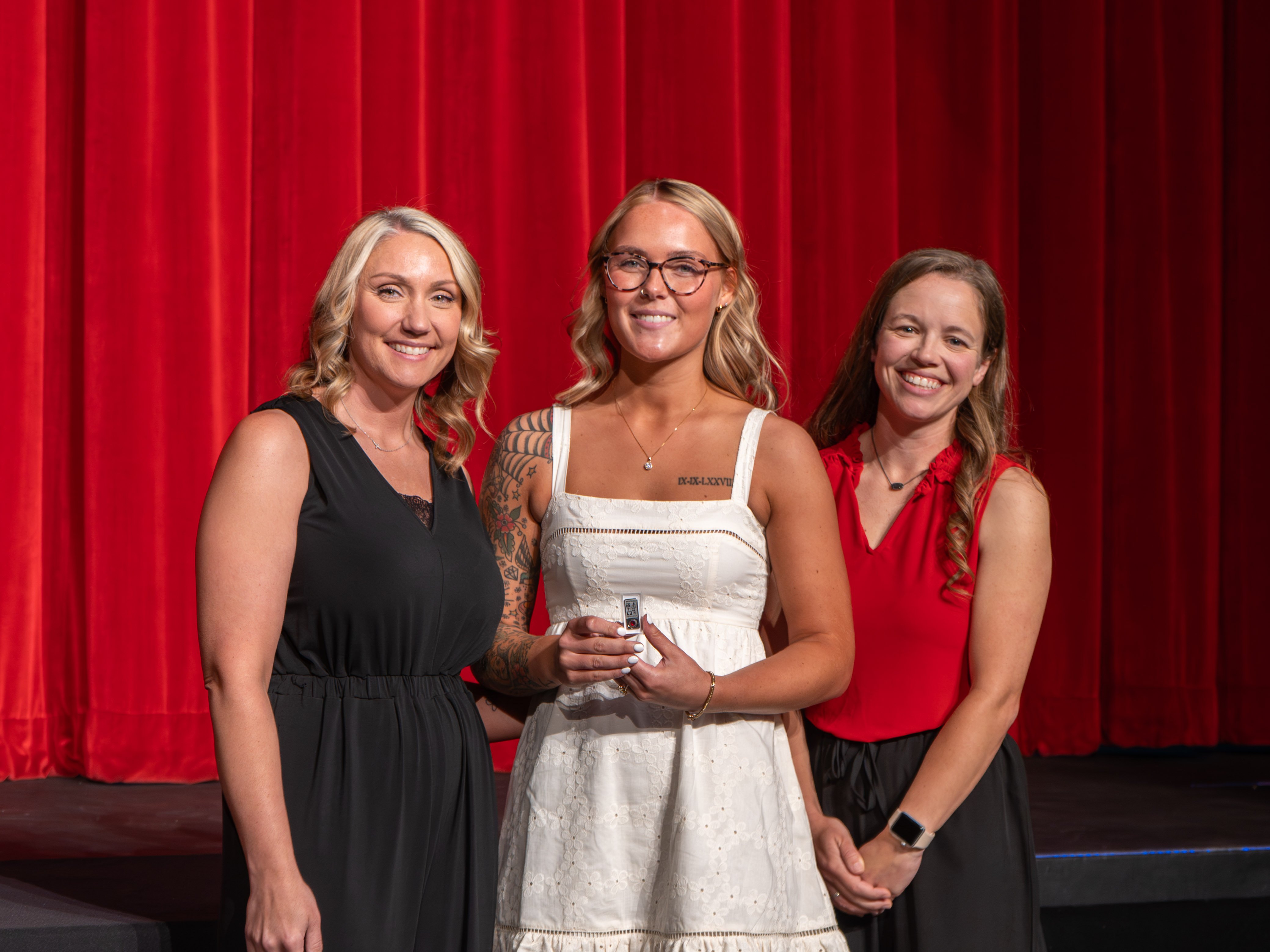 A close-up shot of graduate Chloe Dupuis, a smiling young woman with blonde hair and a shoulder tattoo wearing a white eyelet dress. A male faculty member in a grey polo shirt stands next to her, attaching a program pin to her dress strap against a solid red curtain backdrop.