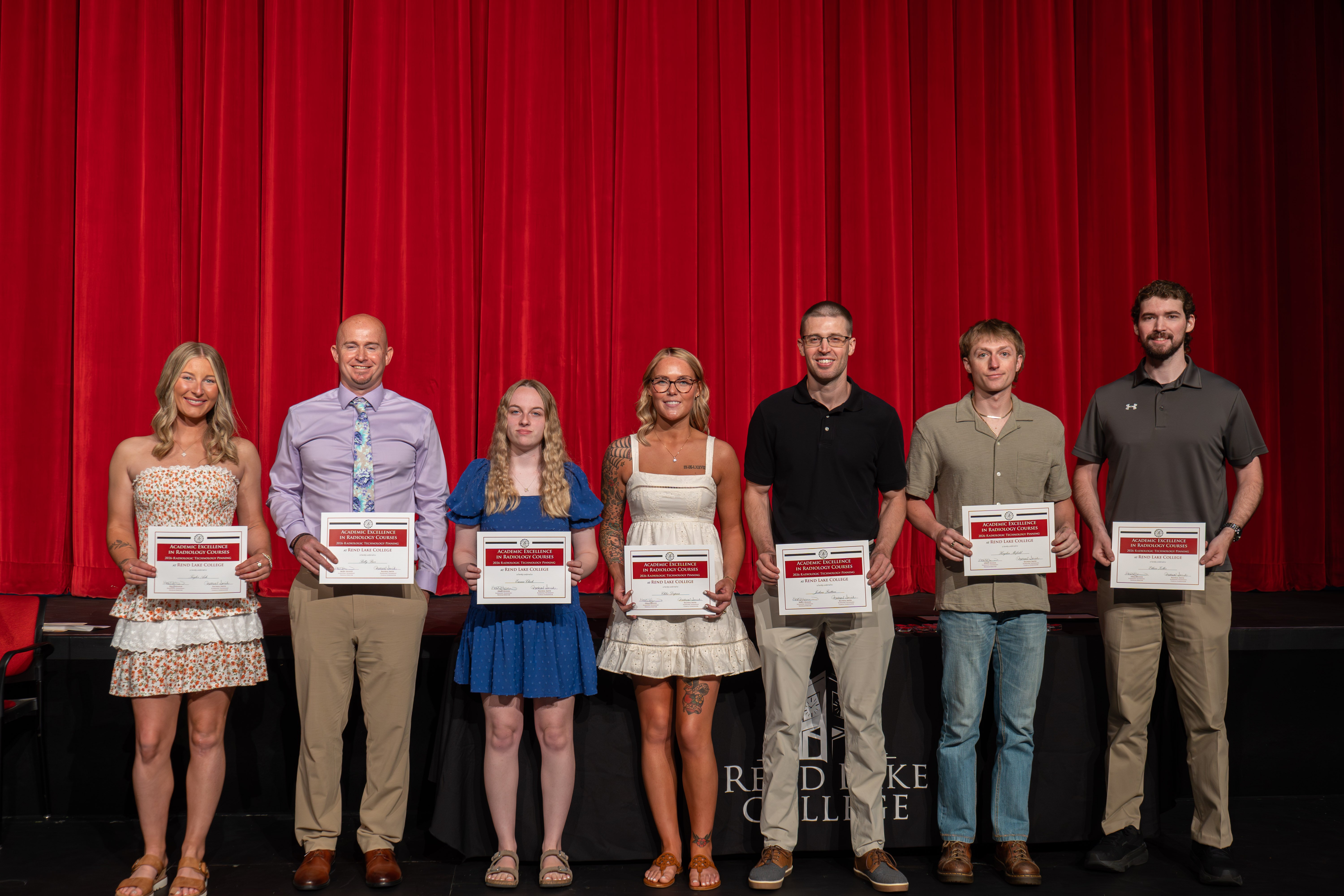 A group portrait of seven Radiologic Technology graduates standing in a line on stage, proudly holding their Academic Excellence award certificates for maintaining a 4.0 GPA. They are positioned in front of a vibrant red stage curtain. From left to right, the graduates are: Taylor Ash in a strapless floral tiered dress; Billy Beer in a light purple dress shirt, floral tie, and khaki trousers; Emma Cluck in a bright blue textured dress; Chloe Dupuis in a white eyelet dress with a shoulder tattoo visible; Joshua Frattini in a black polo shirt and khaki trousers; Hayden Mofield in an olive-green short-sleeved button-up shirt and blue jeans; and Ethan Zoller in a grey Under Armour polo shirt with khaki trousers.