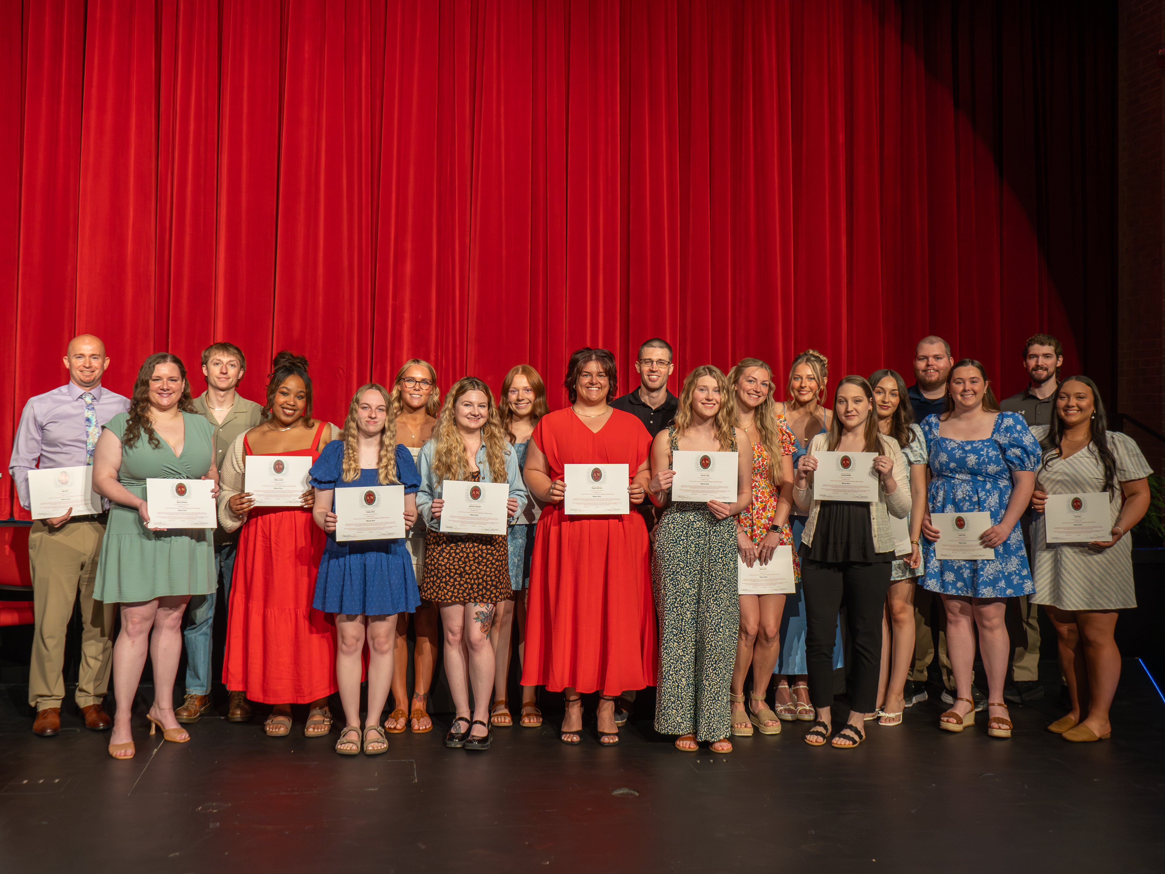 A group portrait of nineteen Lambda Nu national honor society members standing side-by-side in a staggered line on stage. Each student is proudly holding up an honor society certificate. They are posed against a backdrop of a solid, deep red stage curtain. The students are dressed in a mix of colorful semi-formal dresses, skirts, blouses, and button-down shirts with khaki pants, all smiling for the camera.