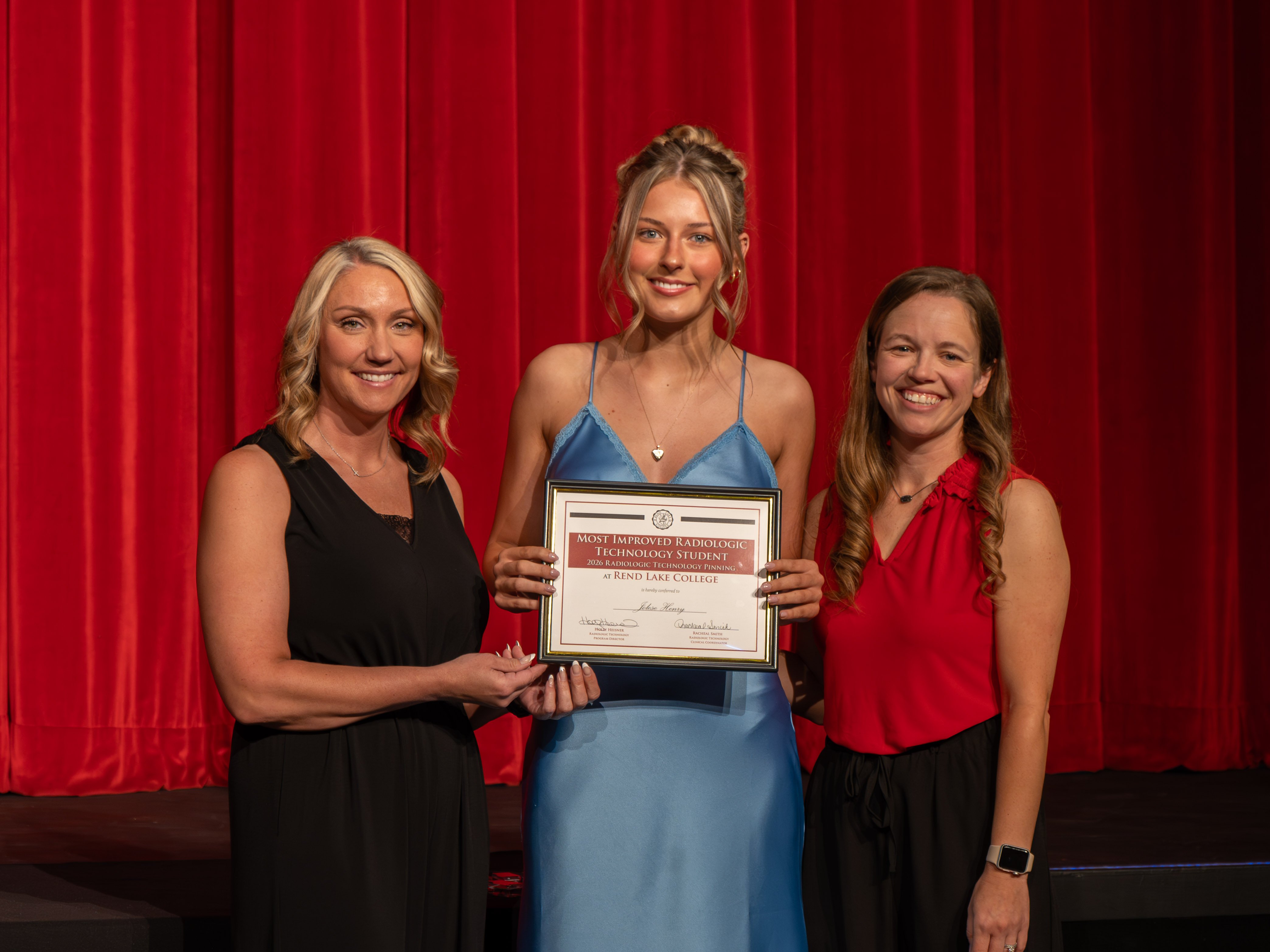 Radiologic Technology faculty member Racheal Smith, wearing a black sleeveless dress, and faculty member Holly Heisner, wearing a red sleeveless blouse and black pants, stand on stage smiling on either side of graduate Jolese Henry. Henry, wearing a blue satin slip dress with her hair styled in an updo, smiles warmly as she holds her framed "Most Improved Radiologic Technology Student" award certificate. A large, vibrant red stage curtain serves as the background.
