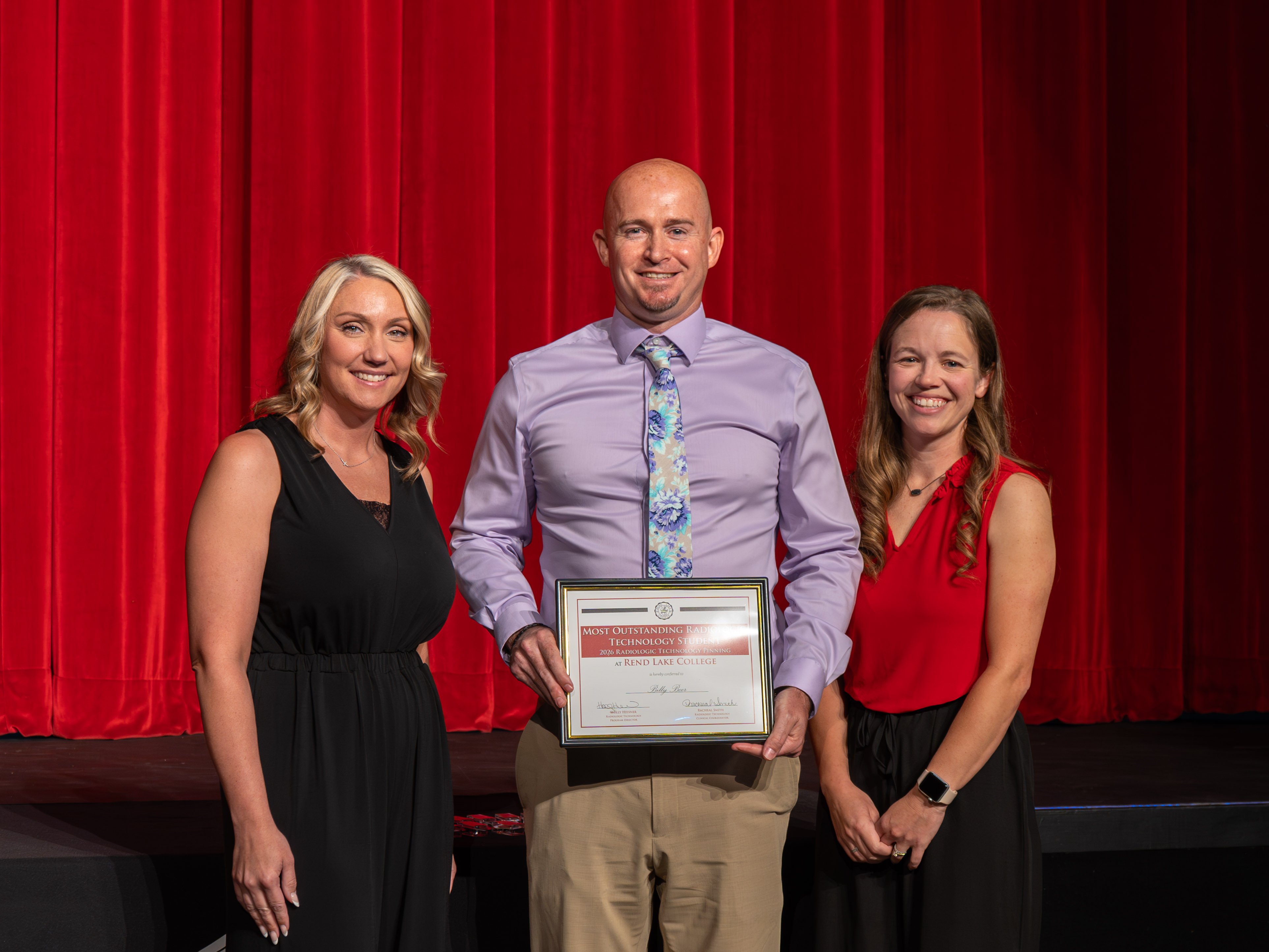 Radiologic Technology faculty member Racheal Smith, wearing a black sleeveless dress, and faculty member Holly Heisner, wearing a red sleeveless blouse and black pants, stand on stage smiling on either side of graduate Billy Beer. Beer, a smiling man with a shaved head, wears a light purple dress shirt, a blue floral tie, and khaki trousers while holding his framed "Most Outstanding Radiologic Technology Student" award certificate. A large, vibrant red stage curtain serves as the solid backdrop.