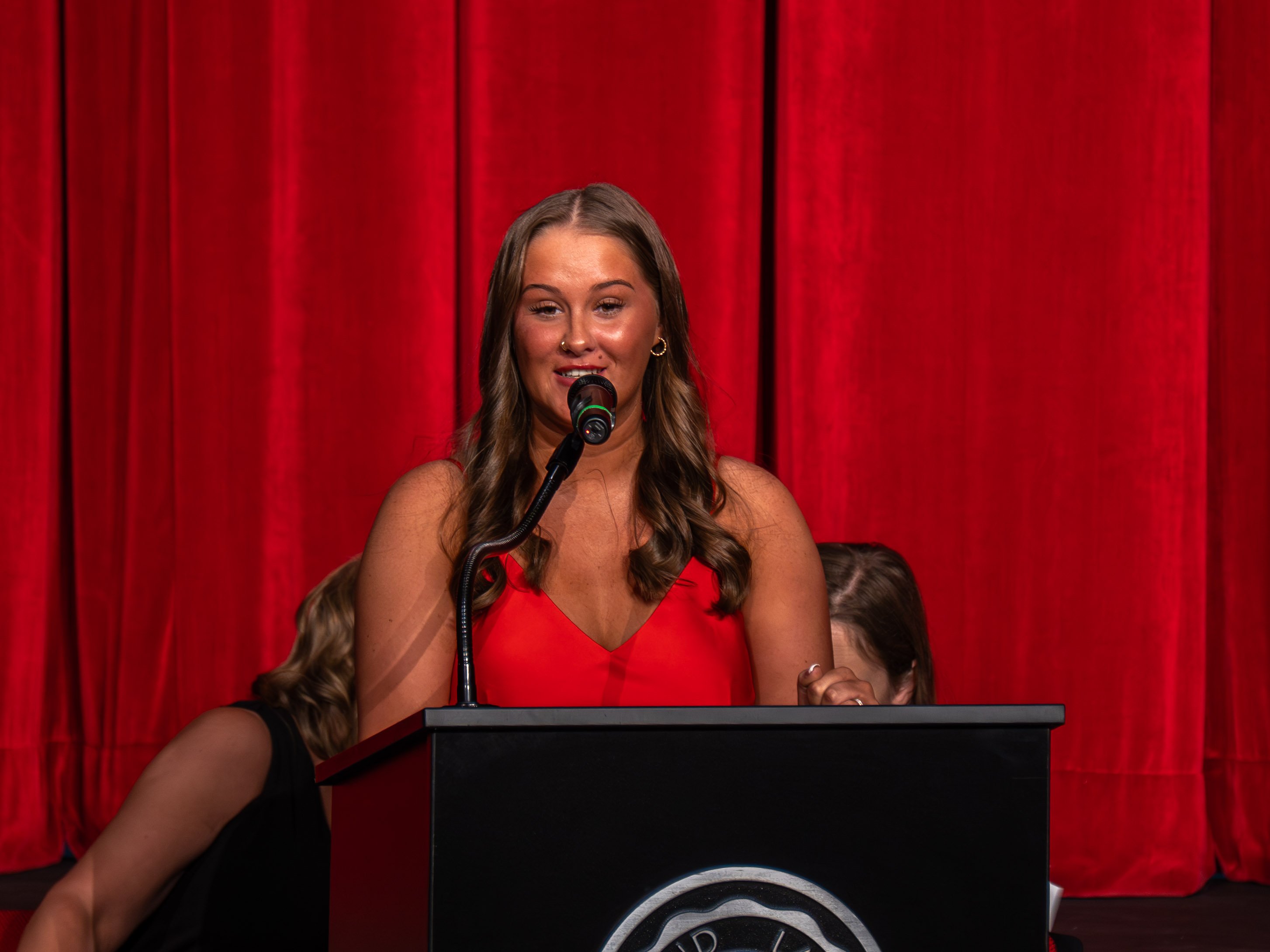 Alt-Text A medium close-up shot of class speaker Maryjane Gentles, a young woman with long, wavy brown hair wearing a red V-neck dress, speaking into a microphone at a black podium during the ceremony. She is smiling warmly while addressing the audience. A vibrant, deep red stage curtain serves as the solid backdrop. The heads of two individuals sitting behind the podium are partially visible on either side, and the top of the Rend Lake College seal can be seen on the front of the podium.