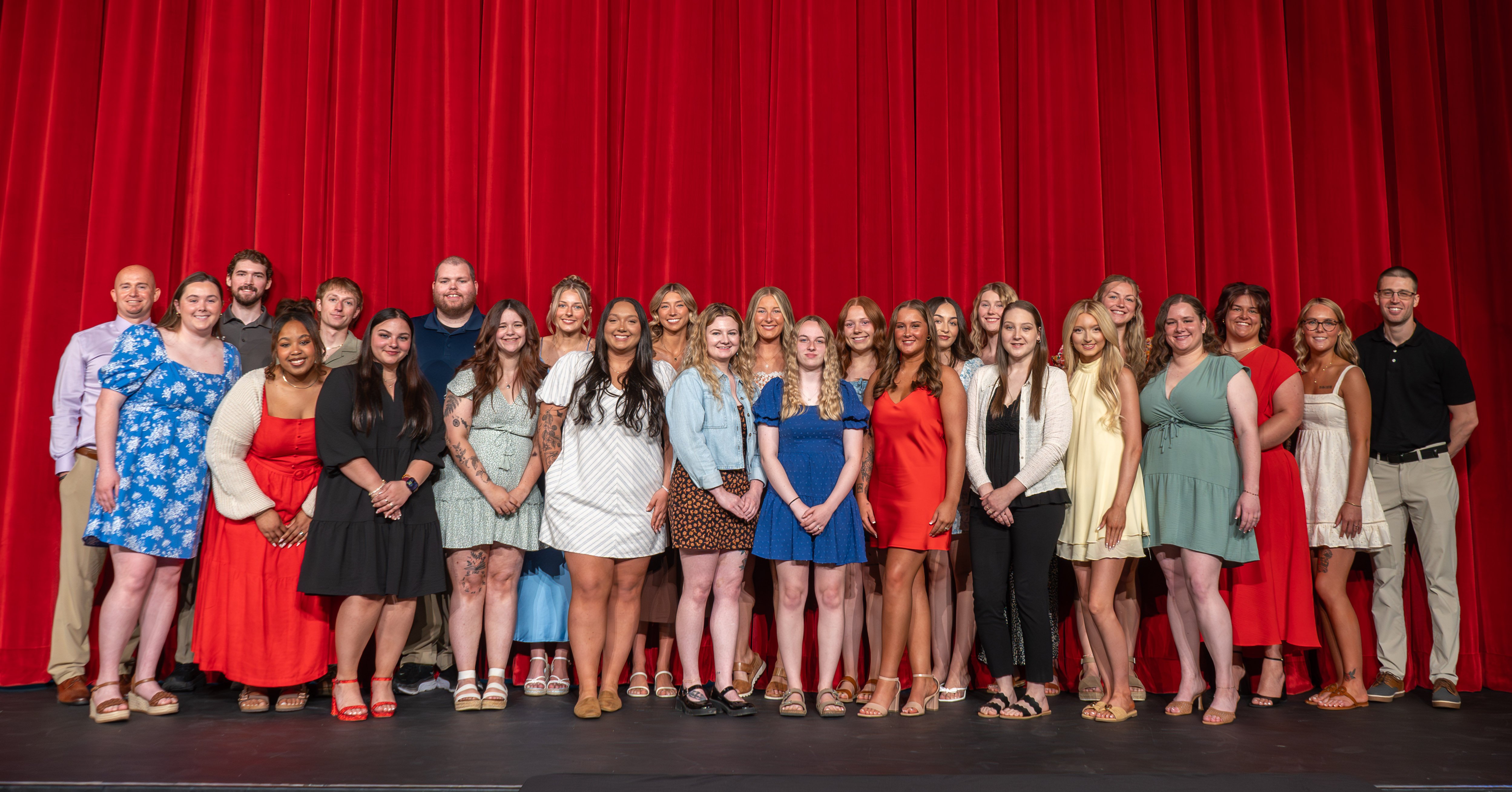 A wide group portrait of twenty-five Radiologic Technology graduates standing in a line on a stage in front of a large, vibrant red curtain. Three male faculty members stand interspersed with the group—one on the far left, one in the back left, and one on the far right. The graduates, a mix of men and women, are dressed in a variety of colorful semi-formal attire, including dresses, blouses, and dress shirts. They are all smiling and facing the camera for the celebratory class photo.