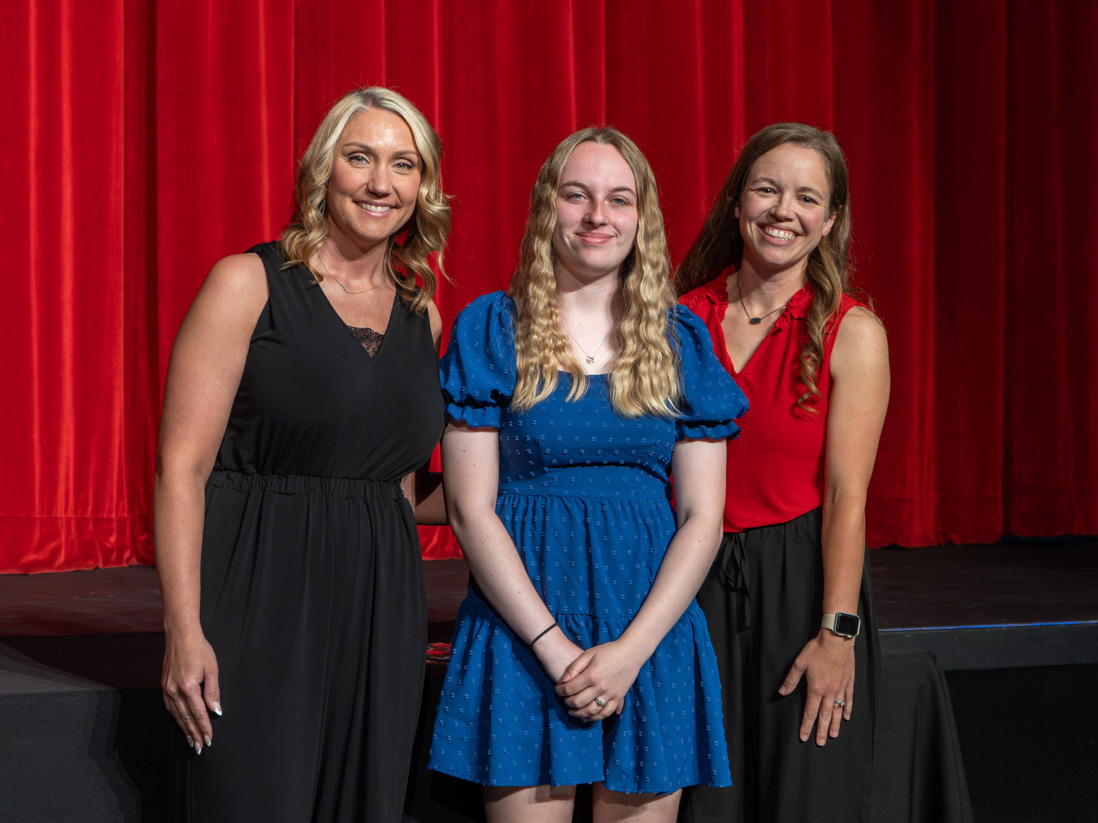 A close-up shot of graduate Emma Cluck, a young woman with long blonde hair wearing a blue textured dress. She smiles warmly as a male faculty member wearing a grey polo shirt stands to her right, carefully fastening a program pin to her dress shoulder. A deep red stage curtain is in the background.