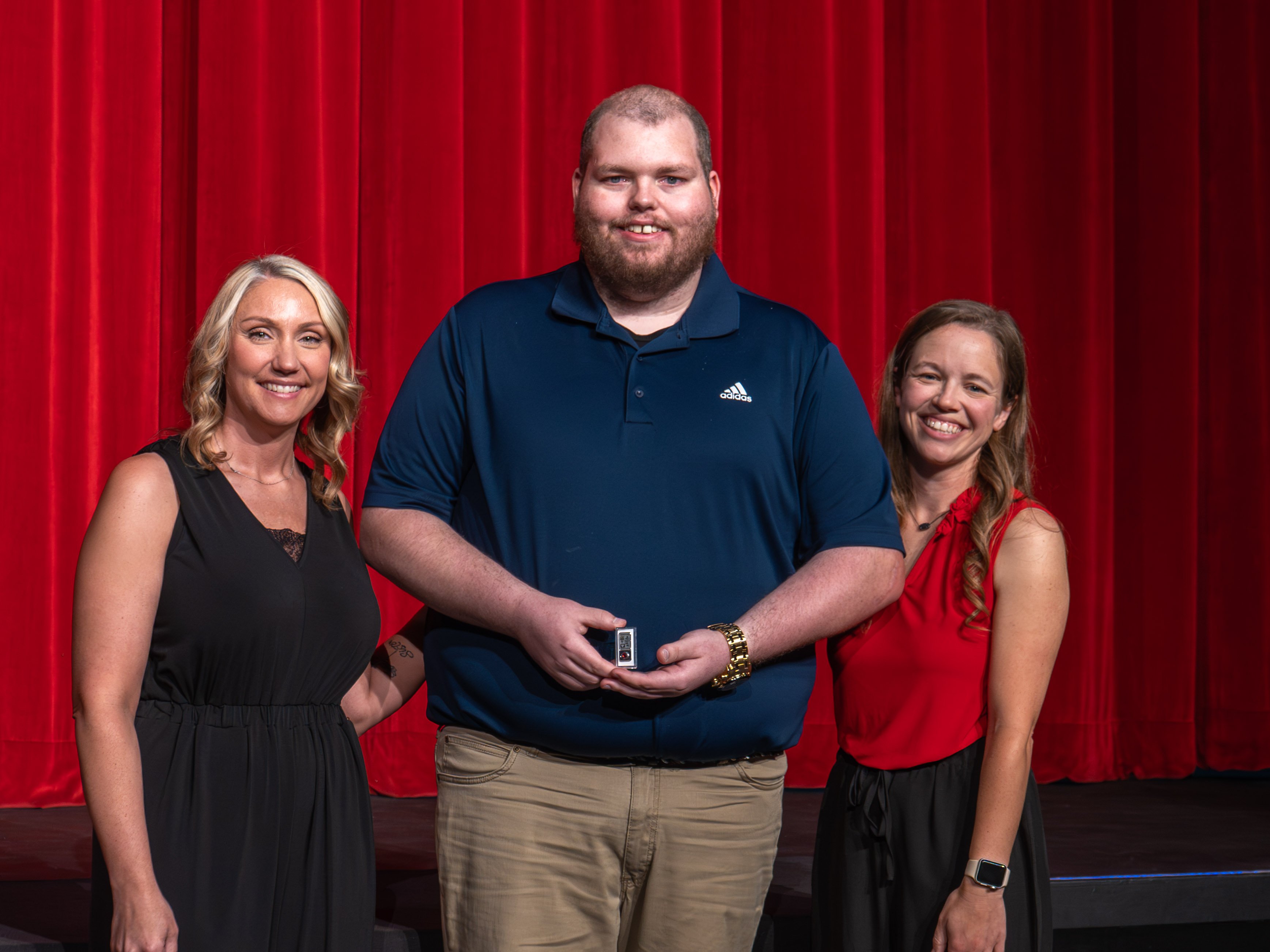A close-up shot of graduate Ethan Smith, a smiling man with a beard wearing a navy blue polo shirt. A male faculty member in a grey polo shirt stands beside him, attaching a program pin near the collar of his shirt. A vibrant red stage curtain serves as the backdrop.