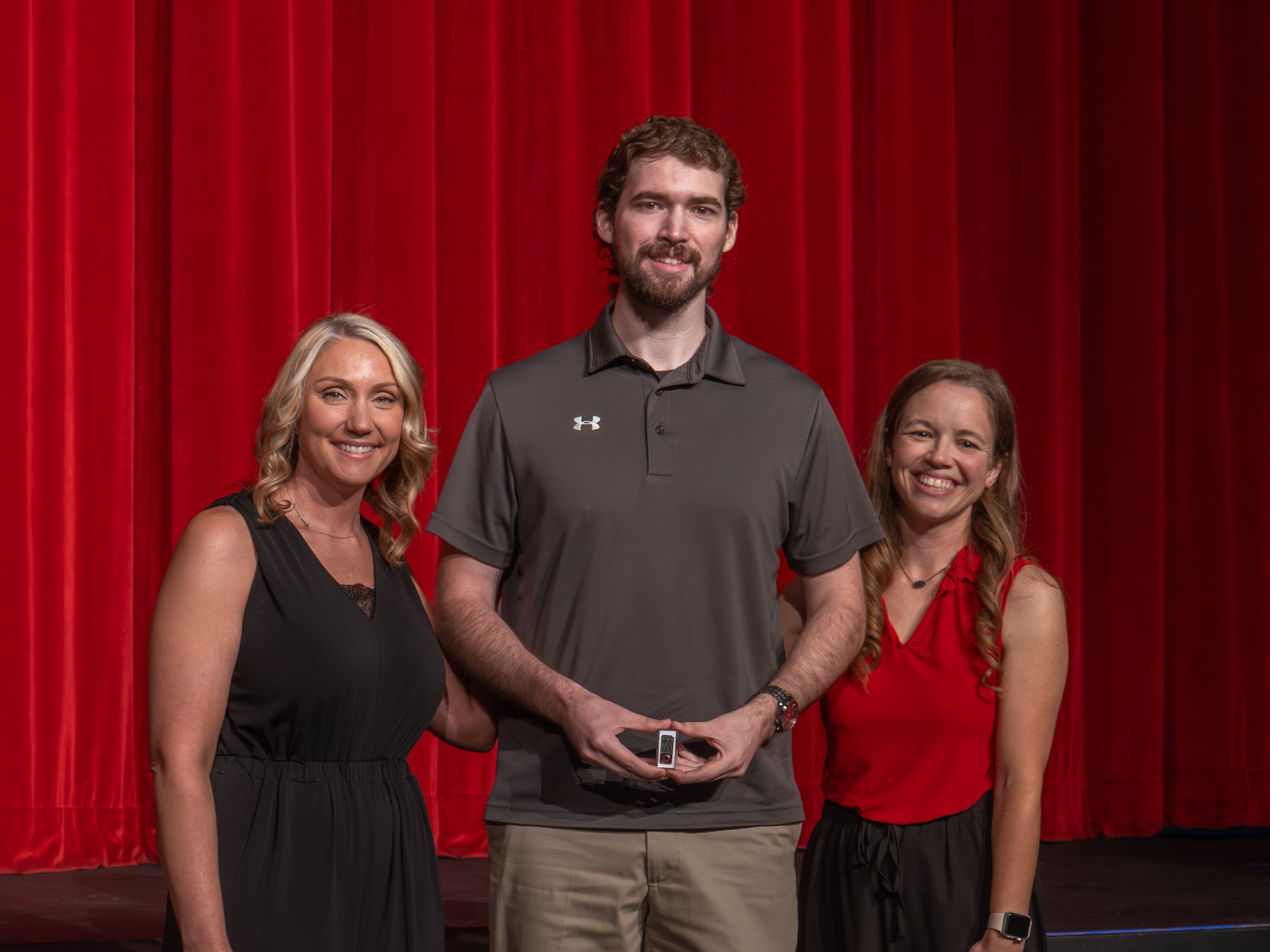 A close-up shot of graduate Ethan Zoller, a smiling man with curly dark hair and a beard wearing a grey polo shirt. Another male faculty member stands to his right, carefully pinning a program emblem onto the front of his shirt against a bright red curtain background.