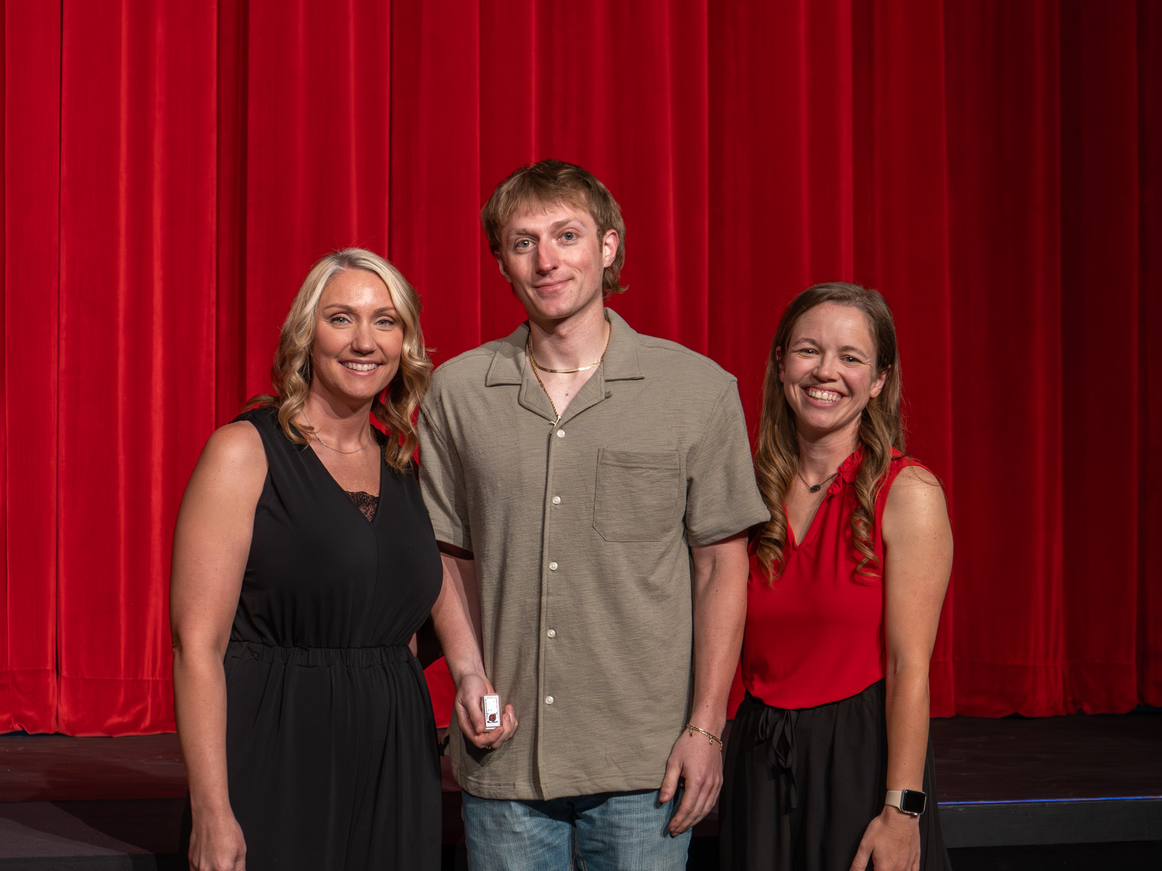 A close-up shot of graduate Hayden Mofield, a smiling young man with short brown hair wearing an olive-green button-up shirt. A male faculty member in a grey polo shirt stands beside him, attaching a program pin to his collar in front of a deep red stage curtain.