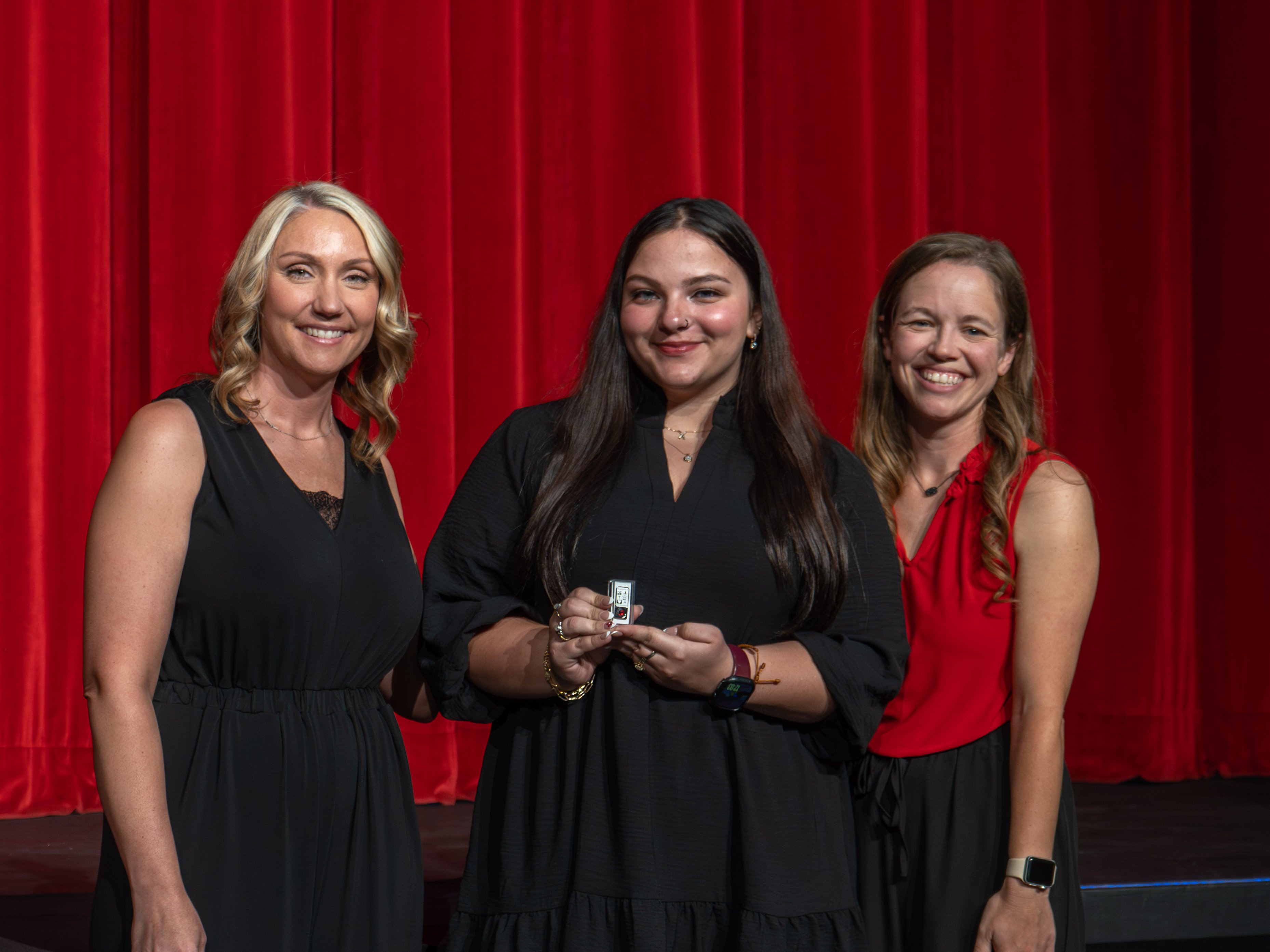 A close-up shot of graduate India Rapach, a smiling young woman with long dark hair wearing a black tiered dress. A male faculty member wearing a grey polo shirt stands to her left, carefully fastening a program pin to her dress collar. A red stage curtain forms the background.