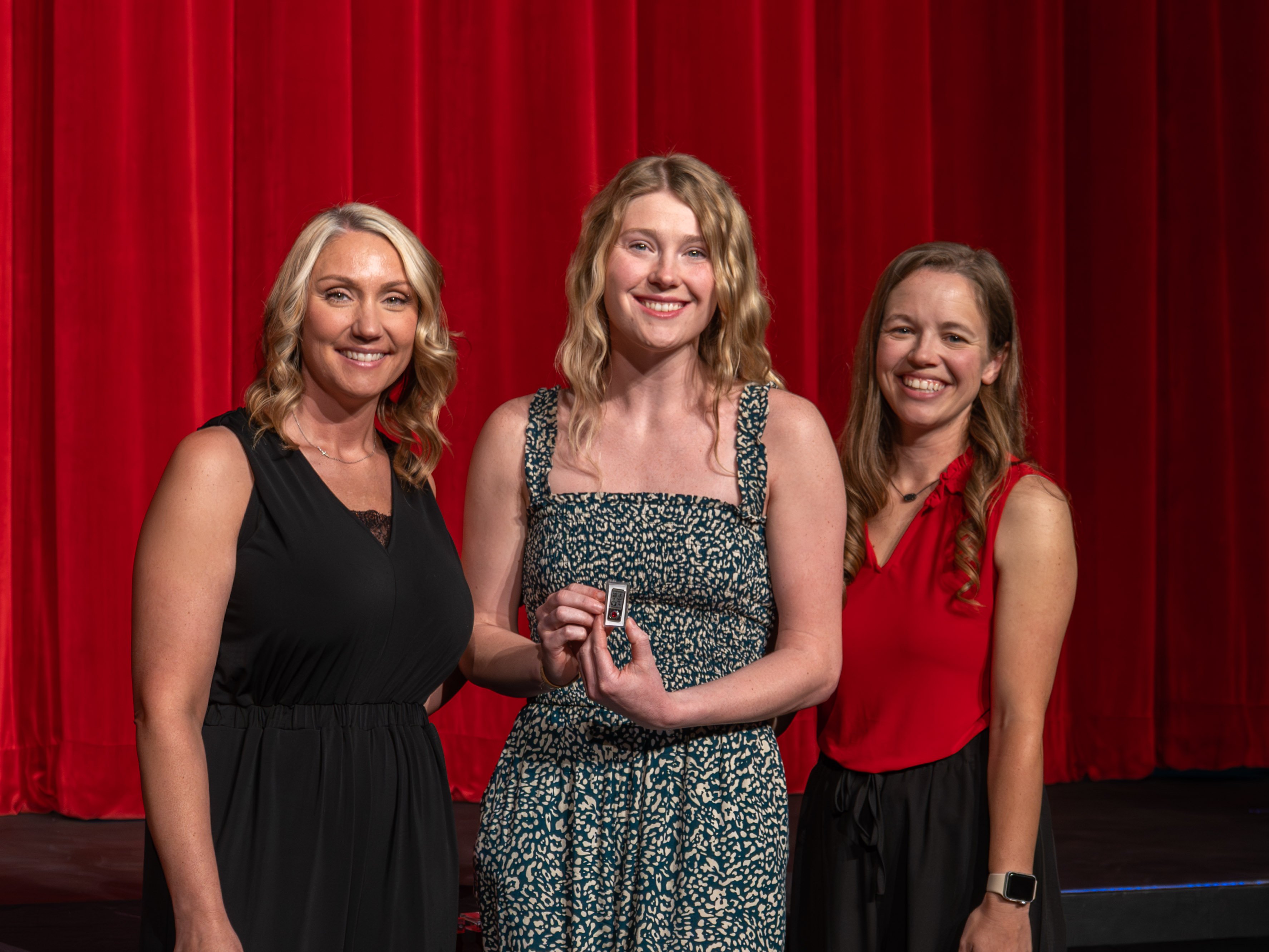 A close-up shot of graduate Janessa Laney, a smiling young woman with wavy blonde hair wearing a green and white patterned sundress. A male faculty member in a grey polo shirt stands beside her, carefully attaching a program pin to the strap of her dress. A deep red stage curtain serves as the background.