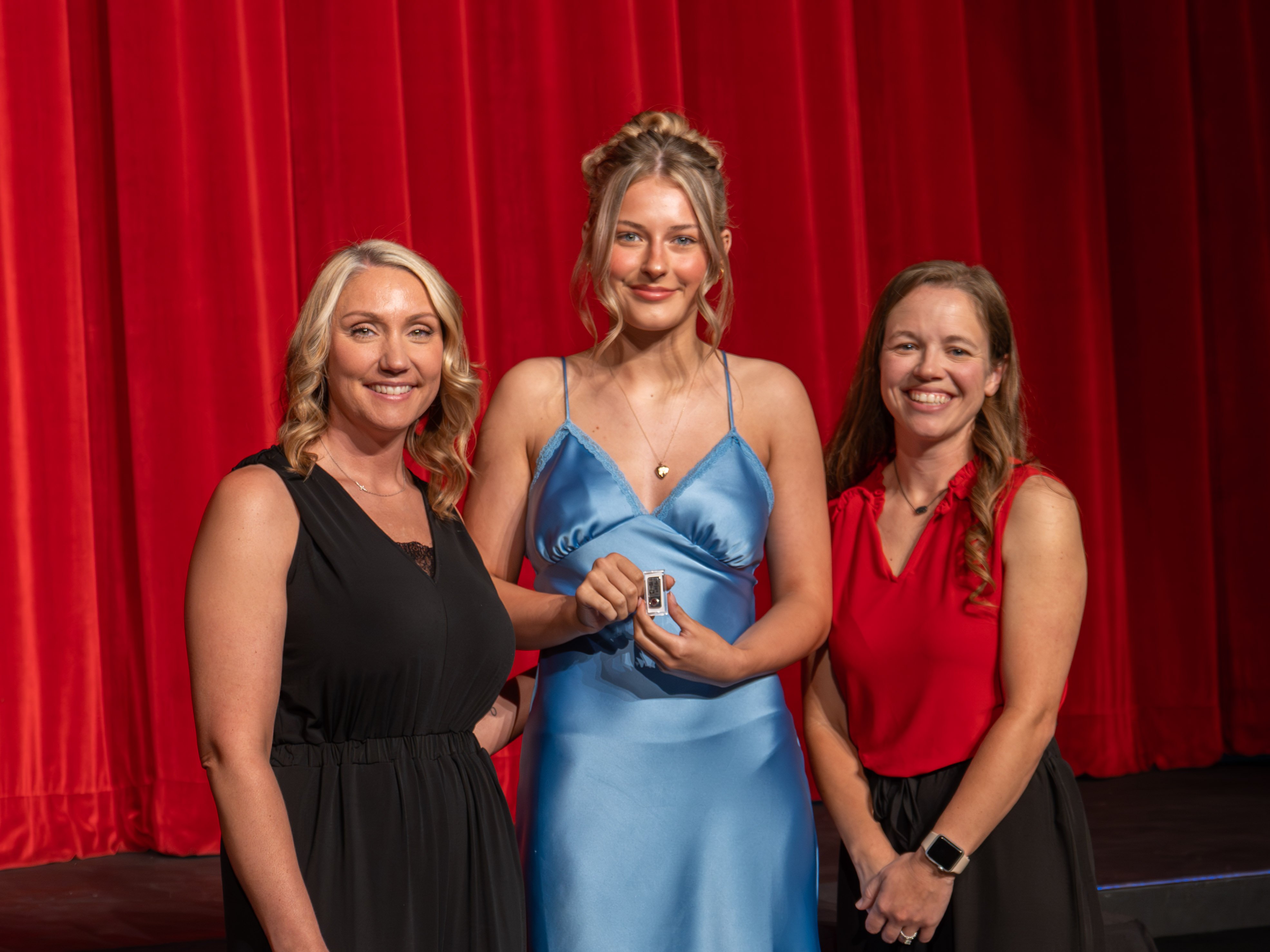 A close-up shot of graduate Jolese Henry, a smiling young woman with her blonde hair in an updo, wearing a light blue satin slip dress. A male faculty member wearing a grey polo shirt stands to her left, placing a program pin onto the strap of her dress against a deep red curtain backdrop.