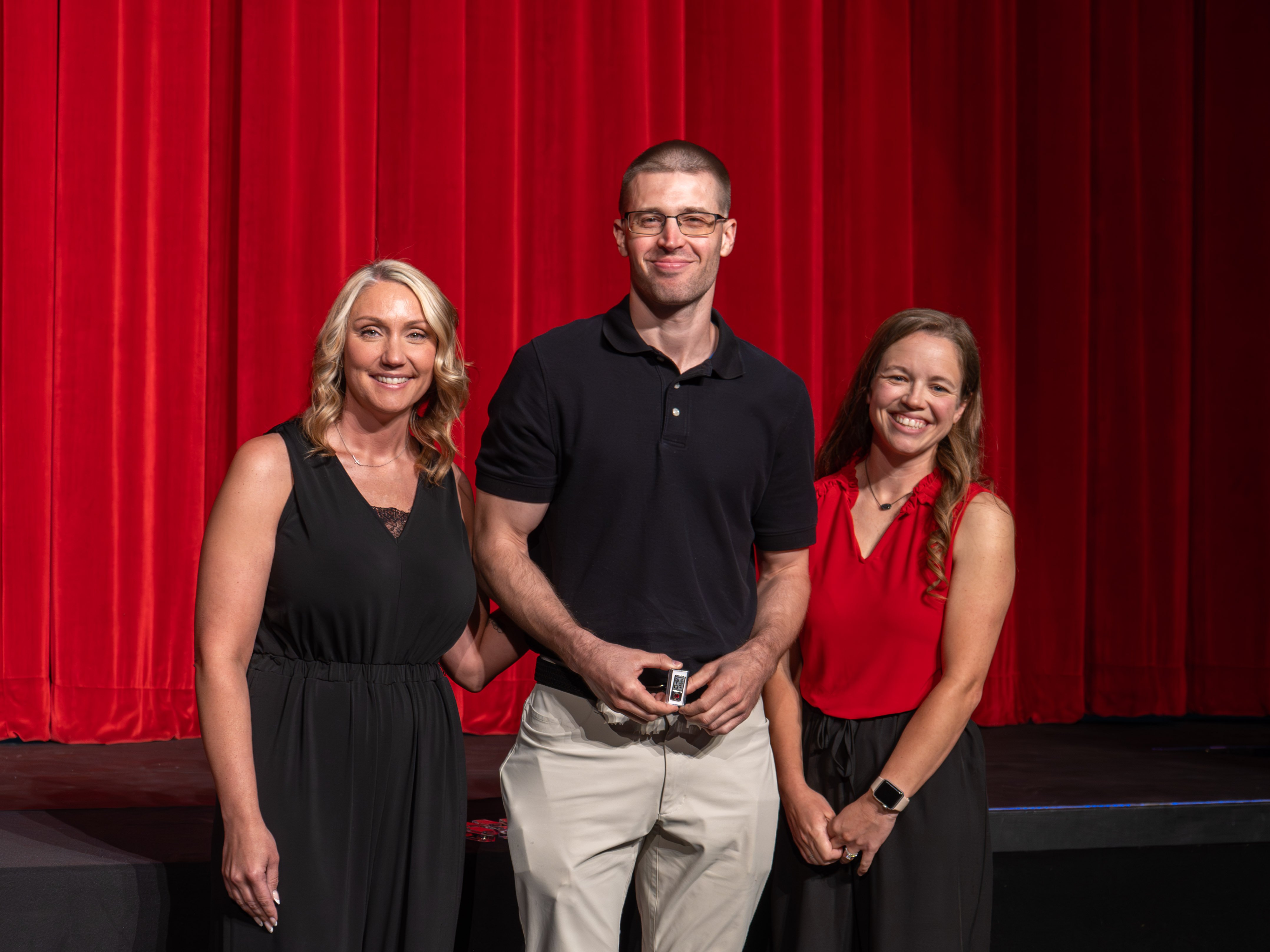 A close-up shot of graduate Joshua Frattini, a smiling man wearing a black polo shirt. A male faculty member in a grey polo shirt stands next to him, carefully fastening a program pin near the collar of his shirt against a bright red stage curtain background.
