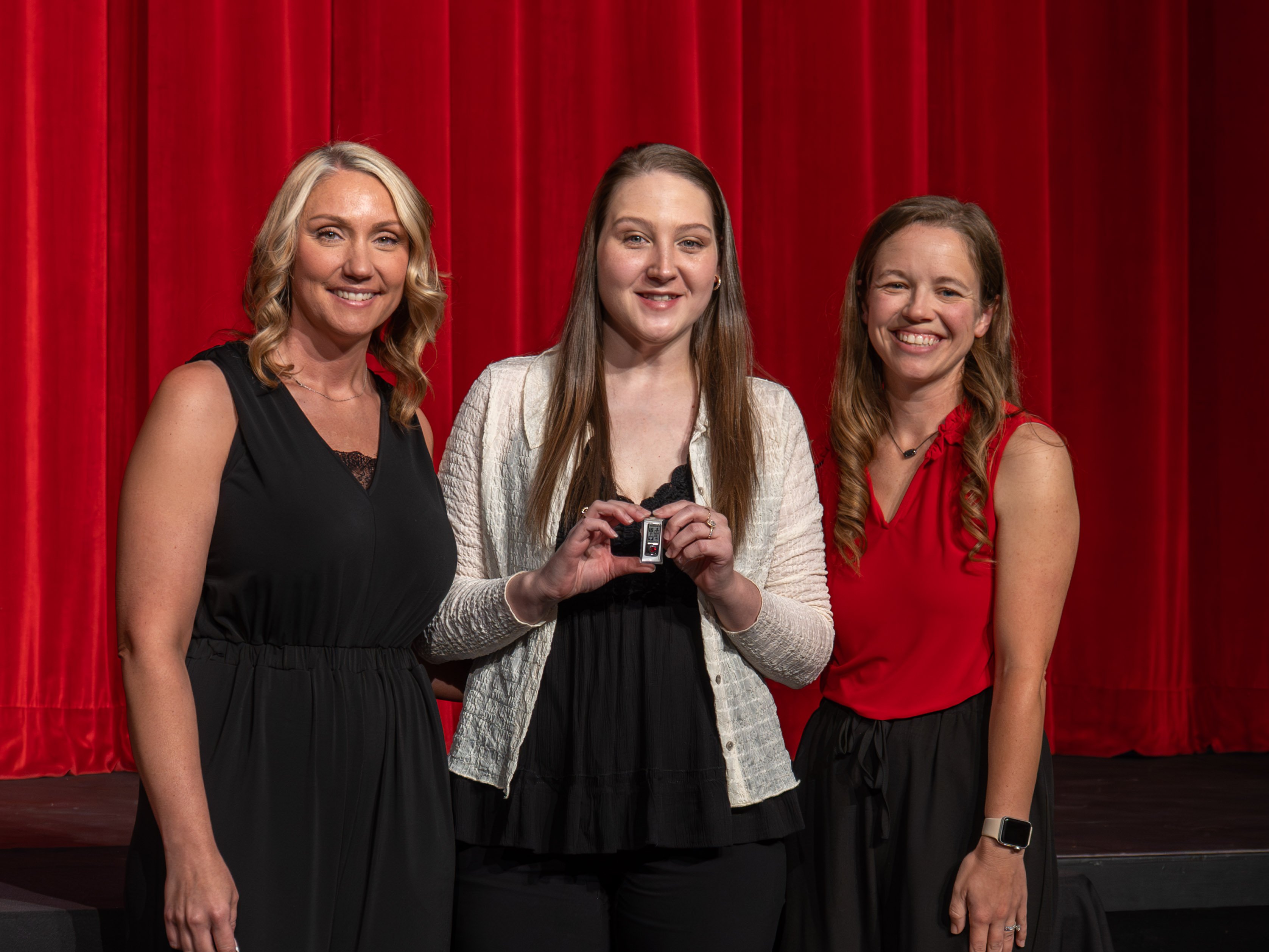A close-up shot of graduate Kassidy Roethler, a smiling young woman with dark wavy hair wearing a black dress with a keyhole neckline. A male faculty member wearing a grey polo shirt stands to her left, attaching a program pin to her dress collar. A red stage curtain forms the backdrop.
