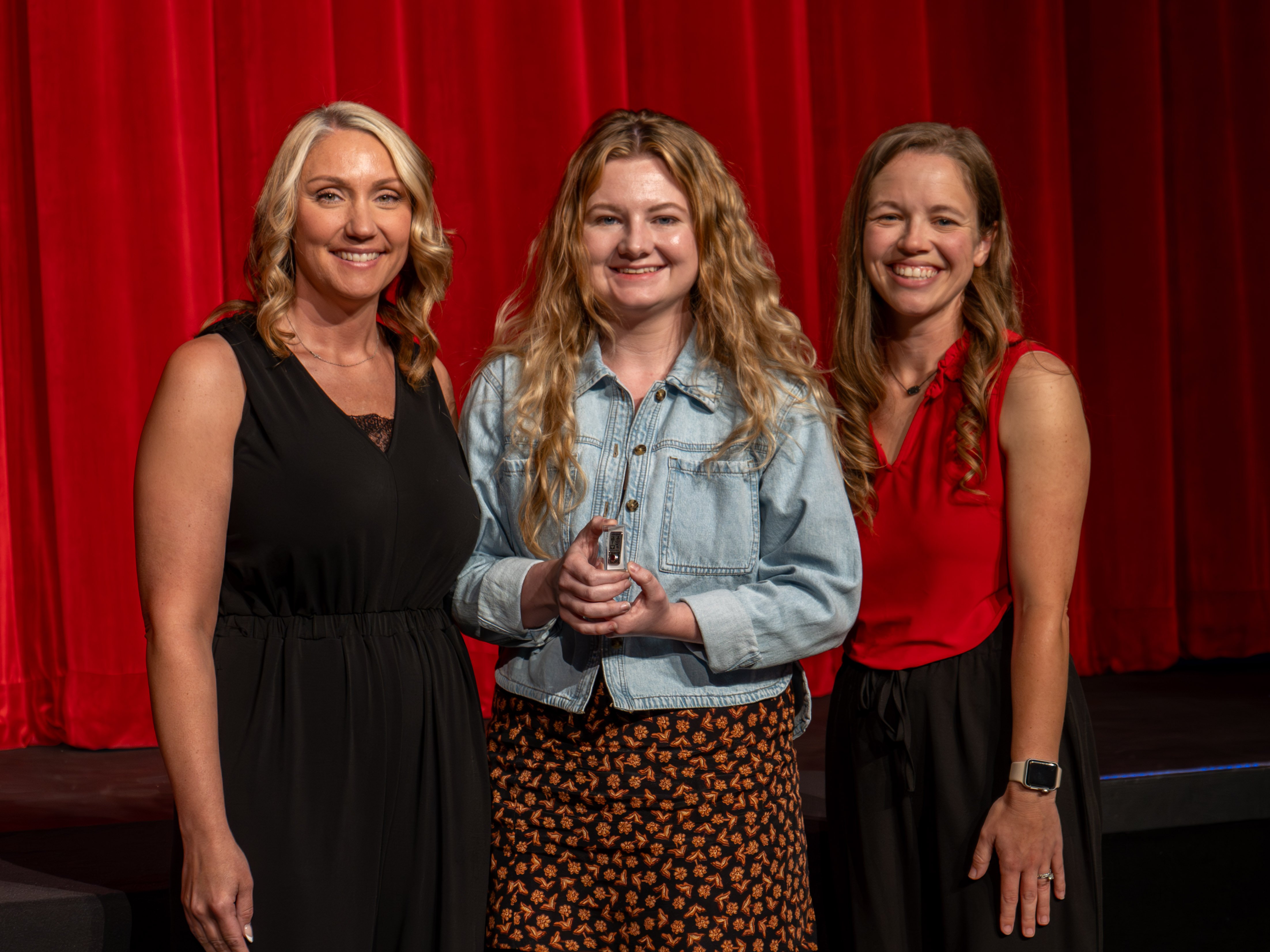 A close-up shot of graduate Kourtney Lipscomb, a smiling young woman with long dark hair wearing a sleeveless cream-colored eyelet dress. A male faculty member in a grey polo shirt stands beside her, attaching a program pin to her dress shoulder strap in front of a deep red stage curtain.