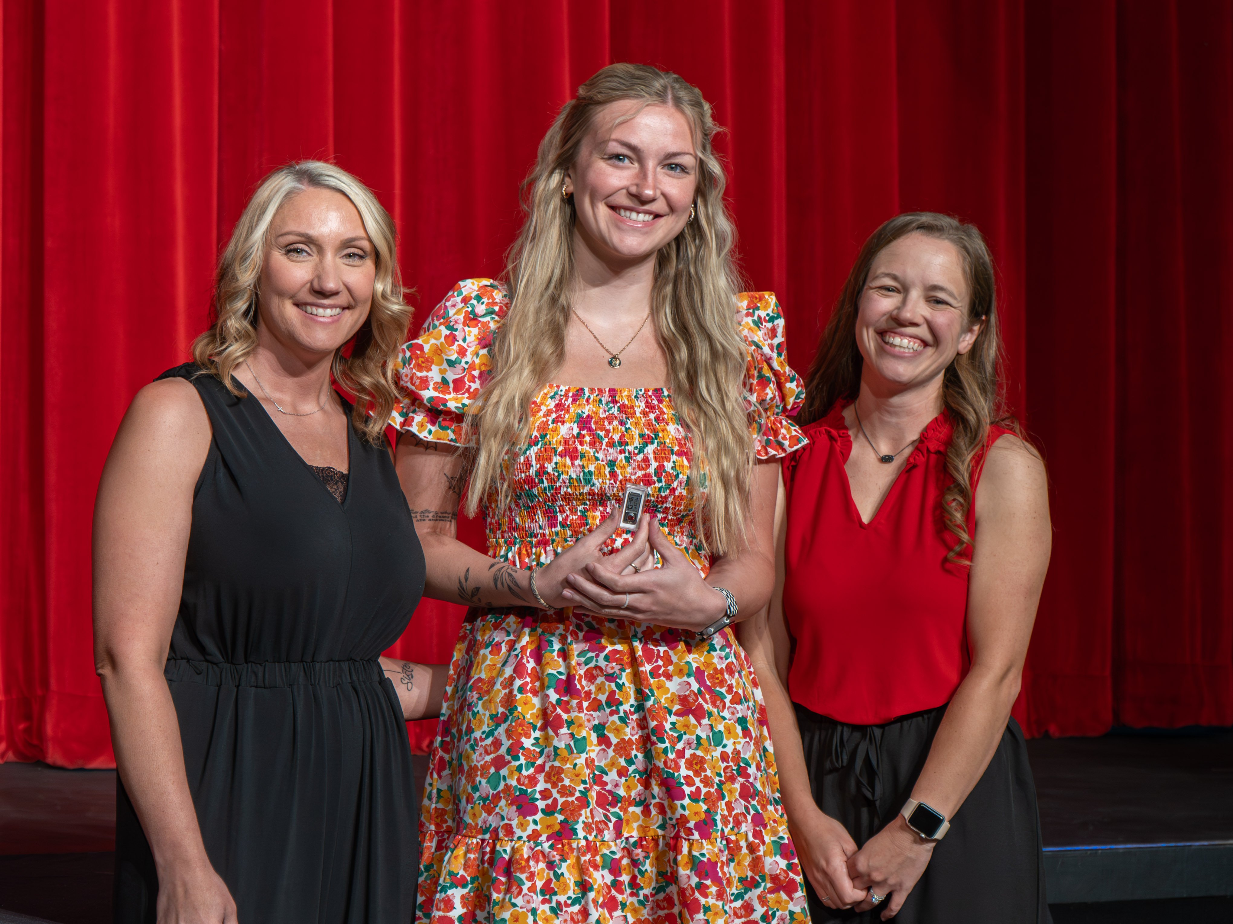 A close-up shot of graduate Kylie Genisio, a smiling young woman with long blonde hair wearing a pastel yellow sleeveless dress. A male faculty member wearing a grey polo shirt stands to her right, carefully fastening a program pin to her dress strap against a solid red curtain backdrop.