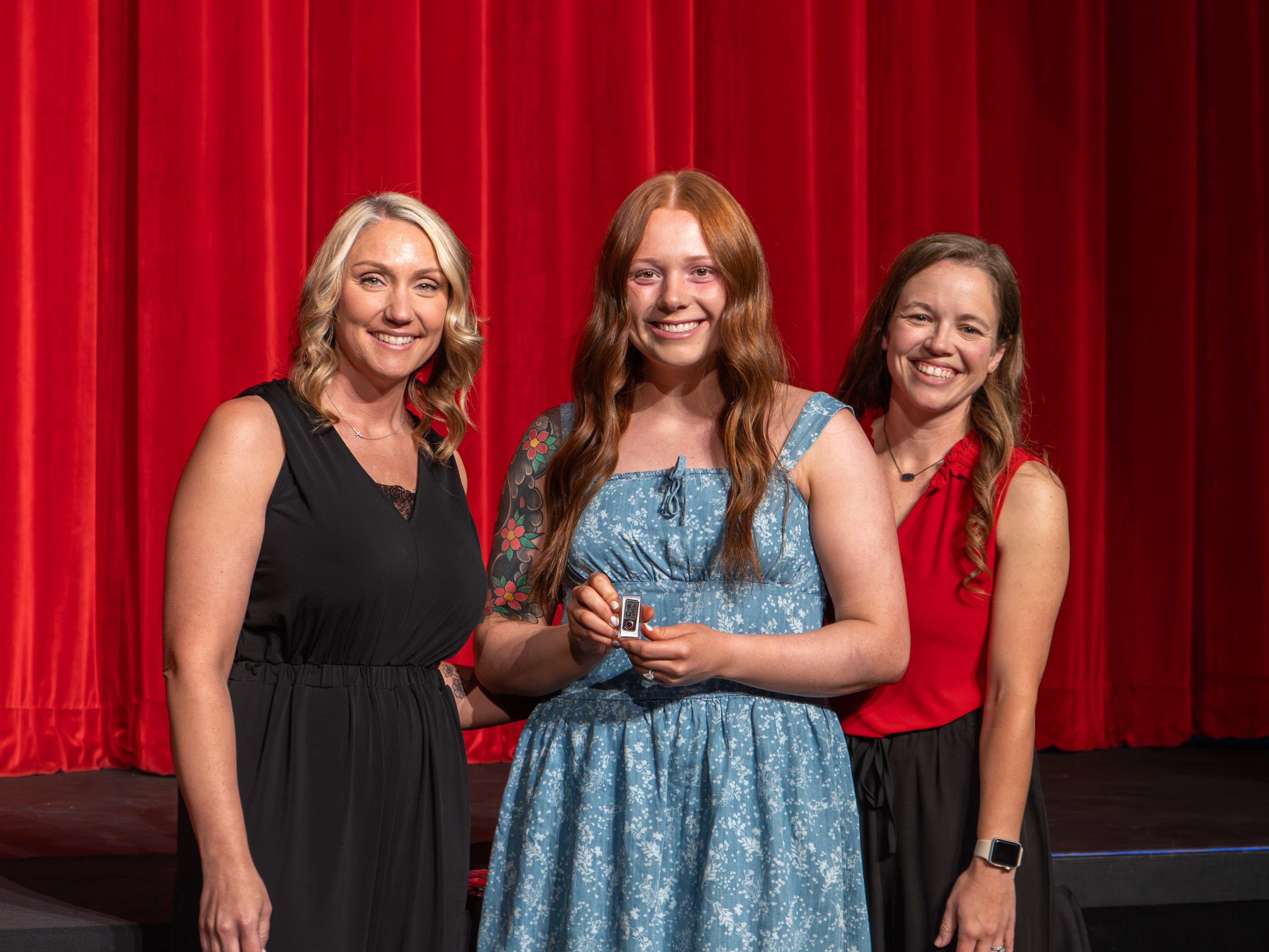 A close-up shot of graduate Makinley Duvall, a smiling young woman with long, wavy red hair wearing a light blue floral dress. A male faculty member in a grey polo shirt stands beside her, carefully attaching a program pin to her dress strap in front of a red stage curtain.