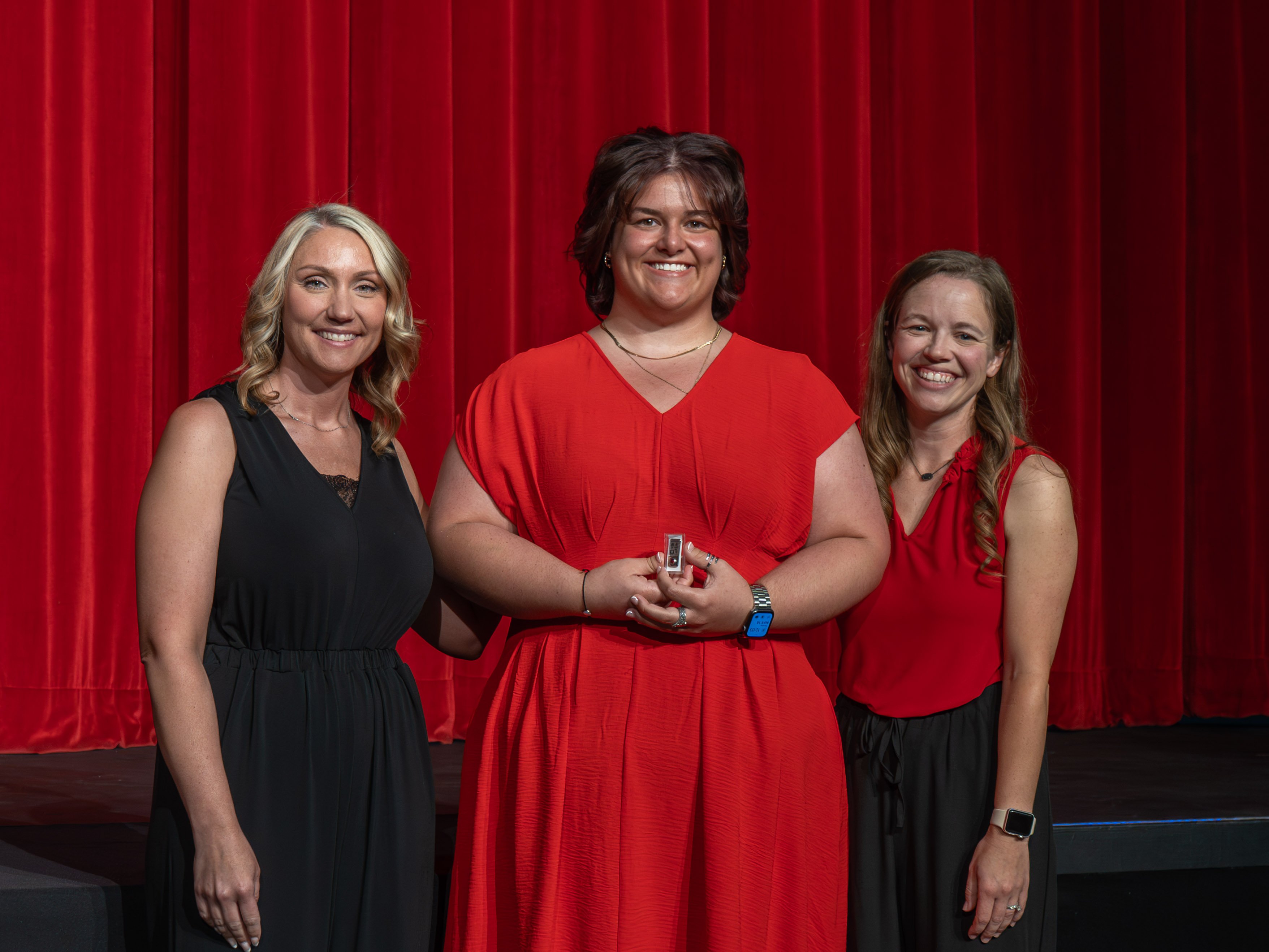 A close-up shot of graduate Morgan Robertson, a smiling young woman with wavy dark hair wearing a light green tiered dress. A male faculty member in a grey polo shirt stands to her left, carefully attaching a program pin to her dress strap in front of a bright red stage curtain.