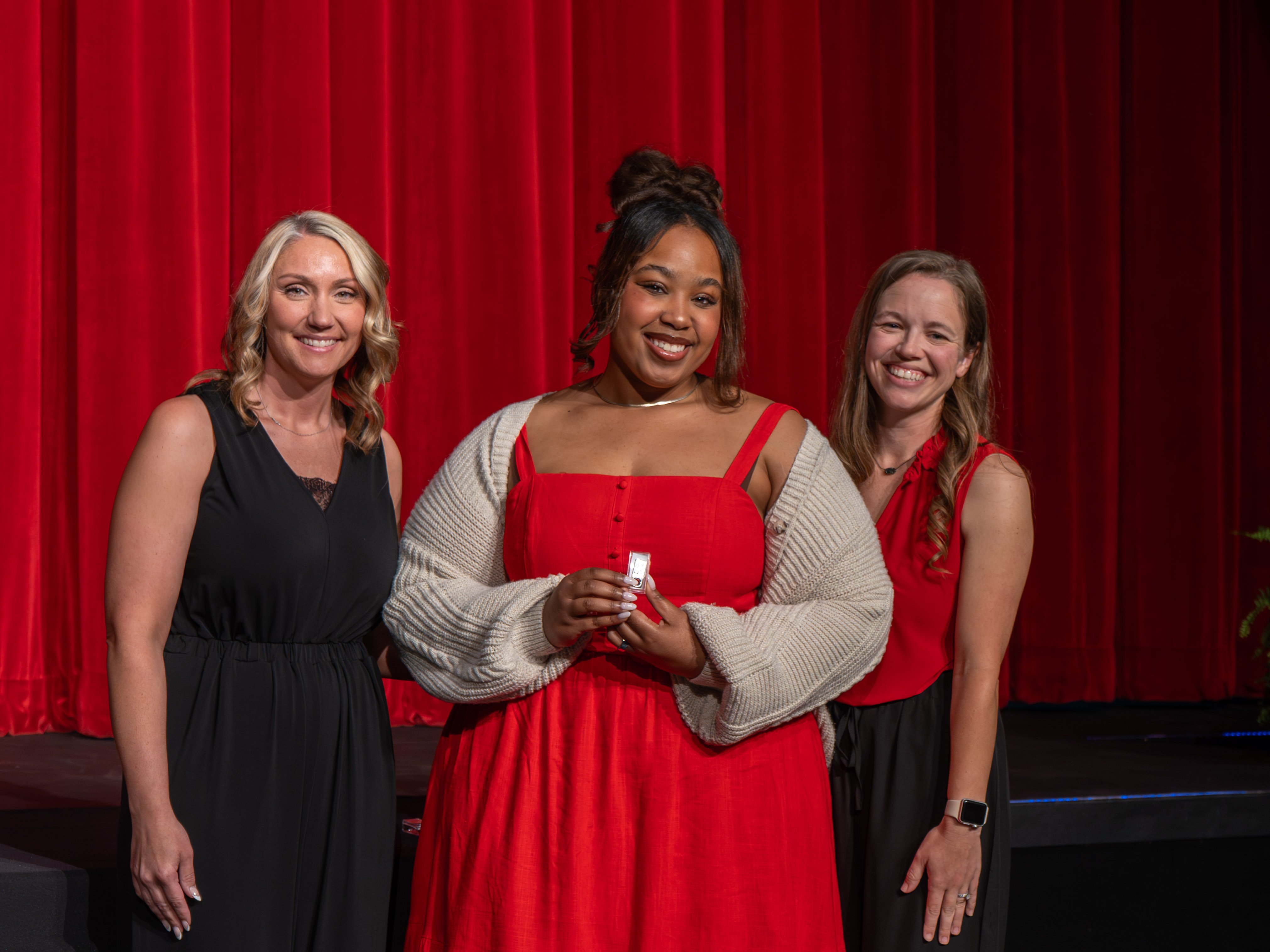 A close-up shot of graduate Nikia Leonard, a smiling woman with glasses and short brown hair wearing a white lace-trimmed sleeveless dress. A male faculty member wearing a grey polo shirt stands beside her, fastening a program pin to her dress strap against a red stage curtain backdrop.