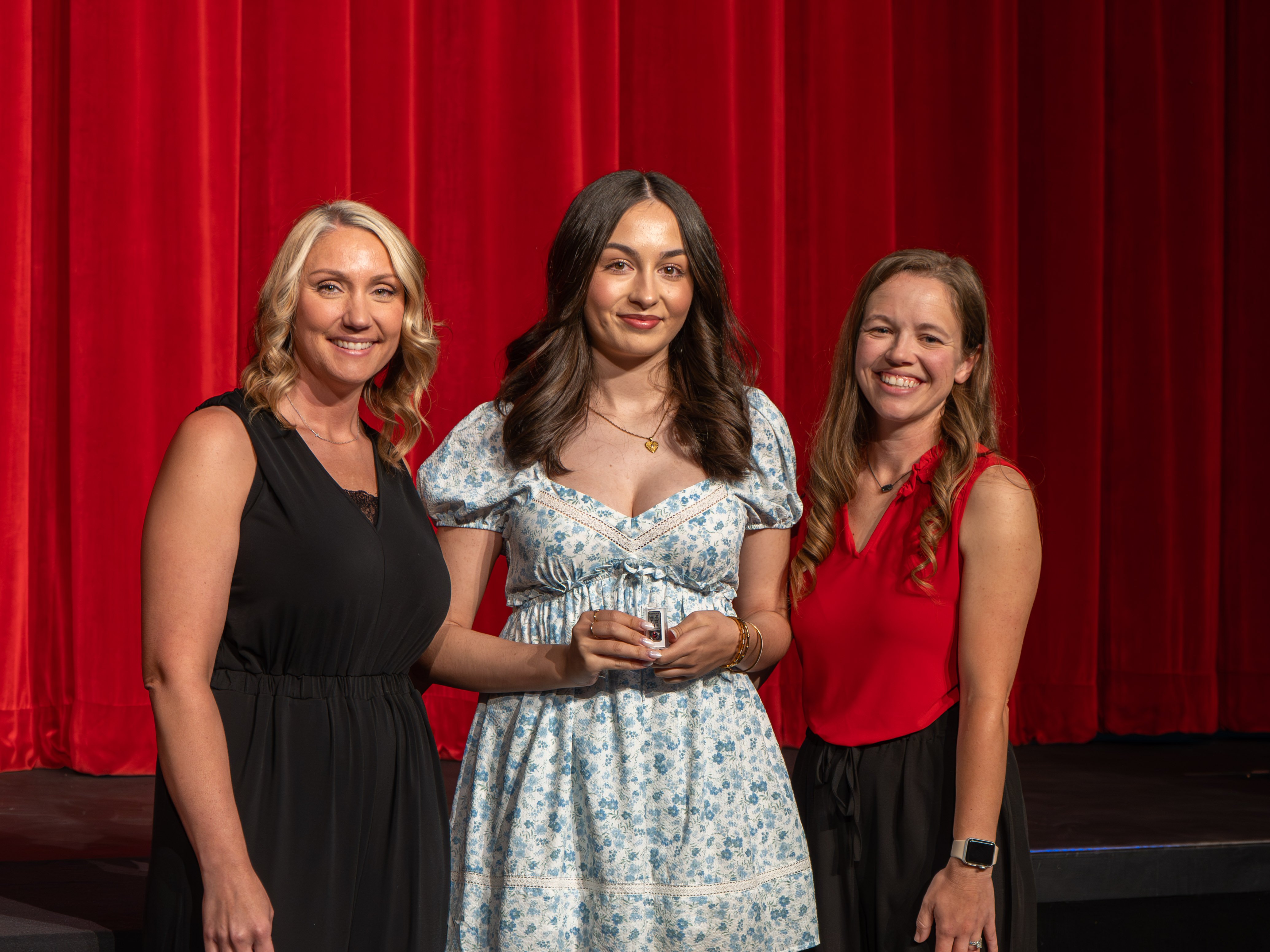 A close-up shot of graduate Noelia Jaque, a smiling young woman with long brown hair wearing a white eyelet lace top. A male faculty member in a grey polo shirt stands to her right, carefully pinning a program emblem near her shoulder against a vibrant red stage curtain background.