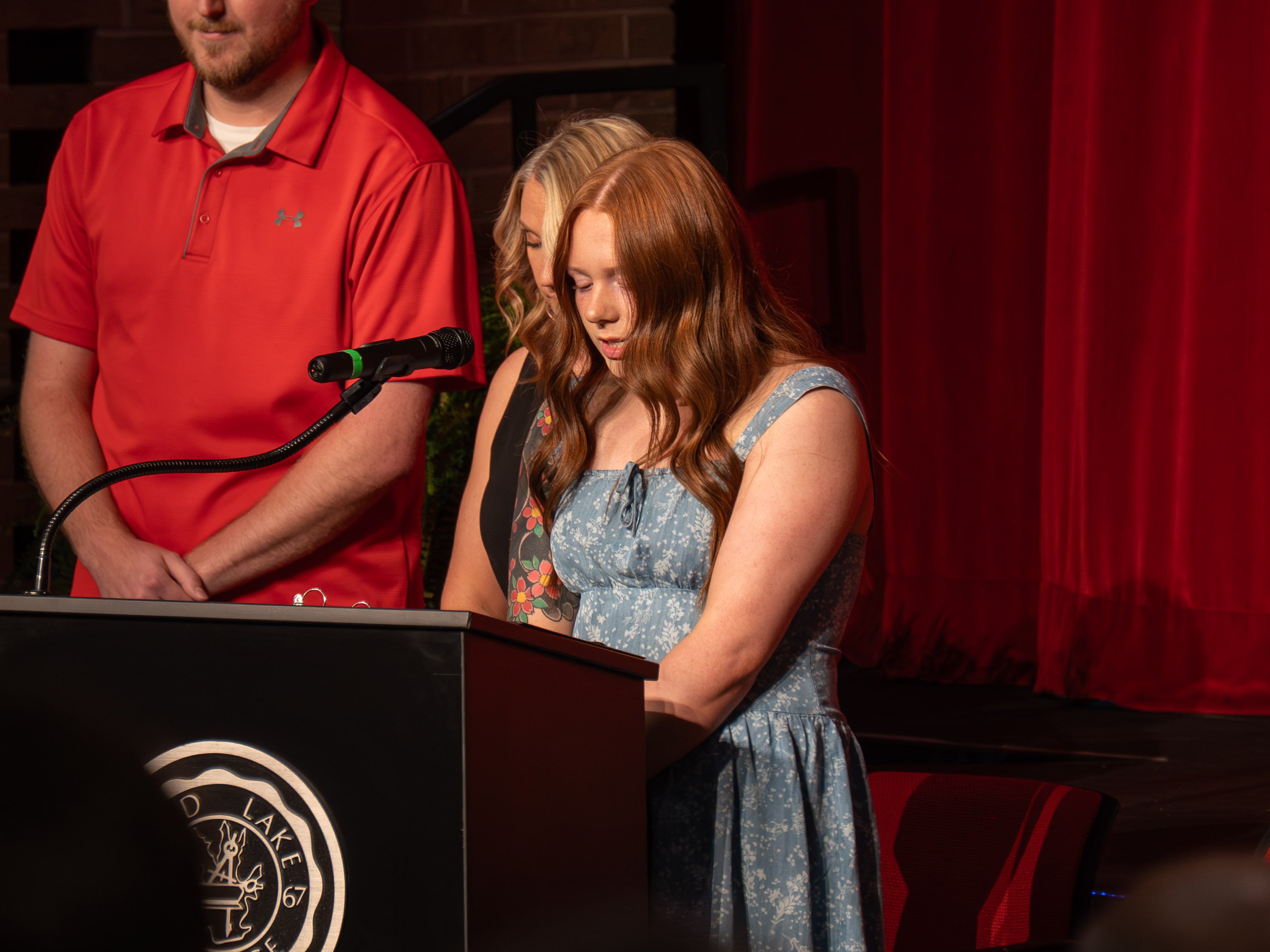 A close-up shot of student Makinley Duvall, a young woman with long, wavy red hair wearing a light blue floral dress, standing at a black podium to deliver a prayer during the pinning ceremony. Her head is bowed and her eyes are closed in focus. In the background, a man in a red Under Armour polo shirt and a woman with blonde hair and a floral arm tattoo stand with their heads bowed in respect. The Rend Lake College seal is partially visible on the front of the podium, and a deep red stage curtain hangs in the background.