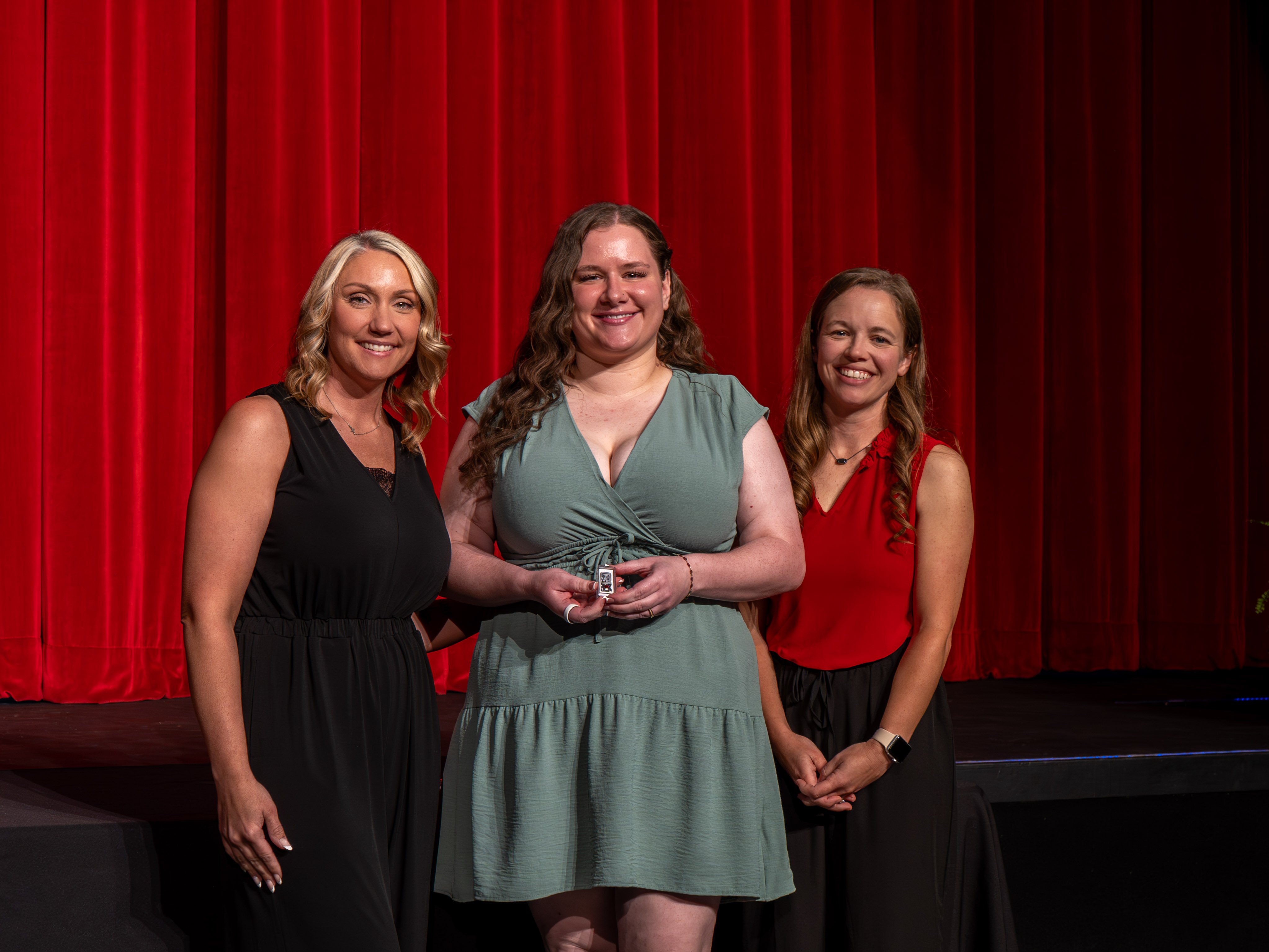 A close-up shot of graduate Renata Bormann, a smiling young woman with long, light-brown hair wearing a blue and white floral dress. A male faculty member wearing a grey polo shirt stands beside her, fastening a program pin to her dress strap in front of a red stage curtain.