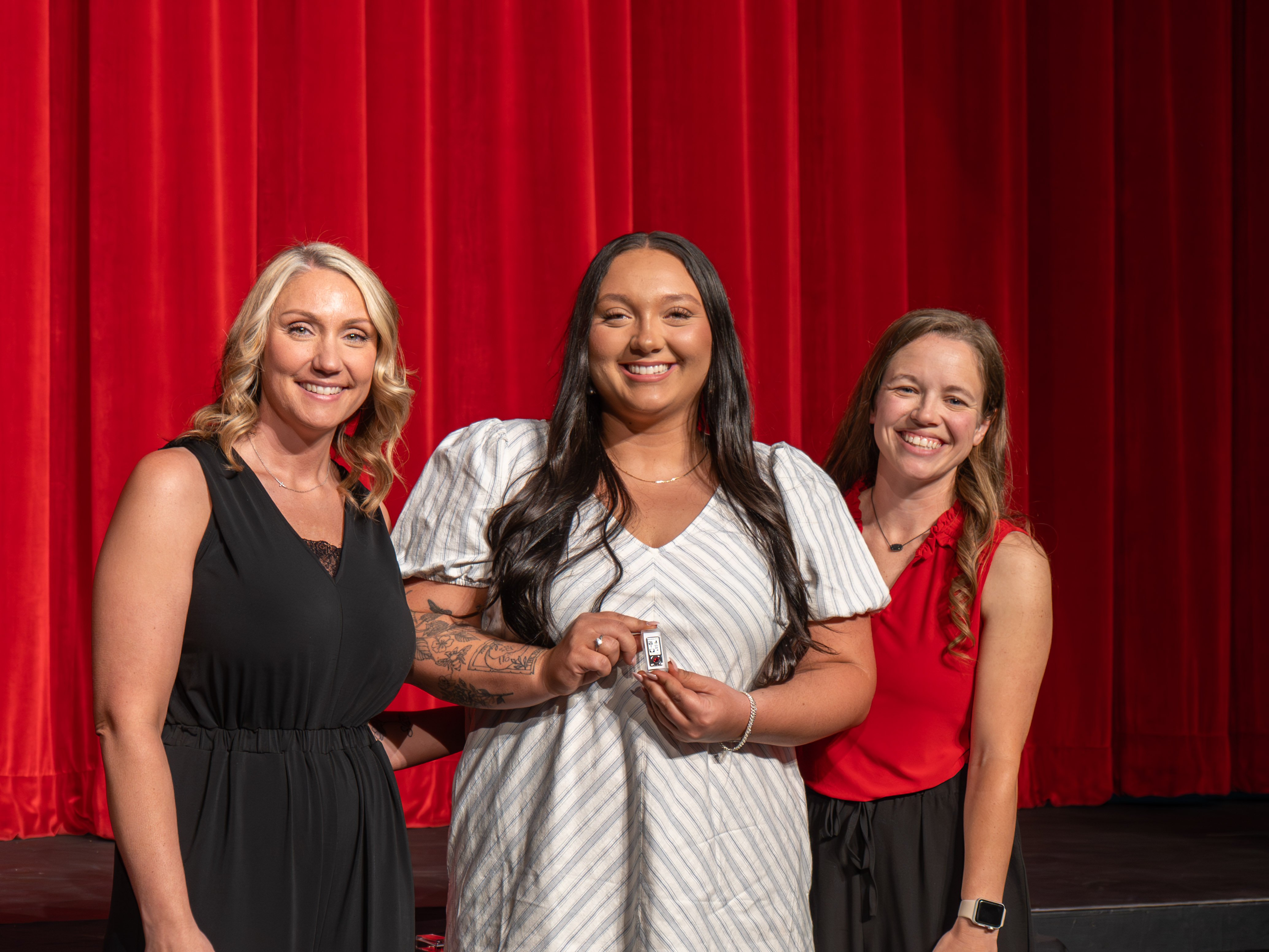 A close-up shot of graduate Tori Evans, a smiling young woman with long blonde hair wearing a bright red sleeveless dress. A male faculty member wearing a grey polo shirt stands beside her, carefully fastening a program pin to her dress shoulder strap in front of a deep red stage curtain.