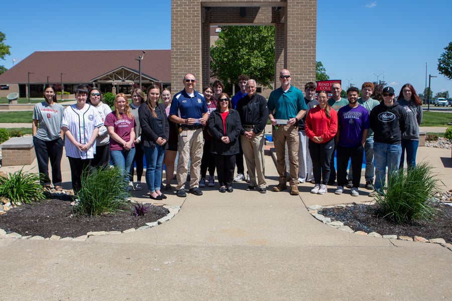 A large group of students and faculty gather in front of the Rend Lake College clock tower on a sunny day. In the center, representatives hold checks to commemorate a donation or award presentation during the 2026 event.