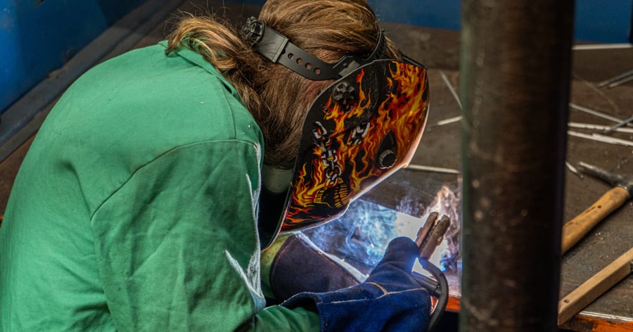 A high school student works on a project during the 2026 Rend Lake College Welding Contest, wearing a green protective jacket and a flame-patterned welding helmet.