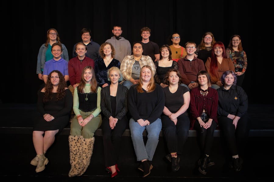 This is a cast photo of the cast of Cabaret. This photo shows a group portrait of 20 individuals, likely the cast of a theatrical production, posed on a stage in front of a solid black curtain. The group is arranged in three distinct rows.  Front Row (Seated on Stage Edge) There are seven people seated in the front row:  On the far left, a person in a black long-sleeved dress and platform sneakers.  A person wearing a light green top, black vest, olive pants, and distinctive fuzzy, spotted boots.  A person in a black pinstripe blazer and red heels.  In the center, a person in a black sweater and blue jeans.  A person in a black t-shirt and black pants.  A person in a maroon button-down shirt and black pants.  On the far right, a person in a black button-down shirt with a small gold emblem on the chest.  Middle Row The middle row consists of seven people standing or sitting slightly elevated behind the first row:  Starting from the left, a person in a bright purple polo shirt.  A person in a dark red long-sleeved shirt.  A person in a dark navy velvet-style top.  In the center, a person in a tan and white plaid flannel shirt.  A person in a black top with a wide neckline.  A person in a maroon sweater.  On the right, a person with long reddish-brown hair in a brown top.  Back Row The back row features six people standing at the rear:  On the far left, a person in a teal shirt and denim jacket.  A person in a dark patterned button-down shirt and glasses.  A person in a grey hoodie.  A person in a plain black t-shirt.  A person in a yellow-orange top with a red strap over their shoulder.  On the far right, two people: one in a black top and one in a floral patterned long-sleeved shirt.