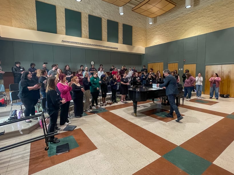 A large mass ensemble composed of multiple community college choirs stands in a rehearsal hall, joining together for a final performance. The diverse group of singers is spread across a multi-level stage and floor area, all facing a grand piano. Director Dr. Christopher Eads, dressed in a dark suit, stands at the piano leading the choir as they perform "Come and Go (To That Land)."
