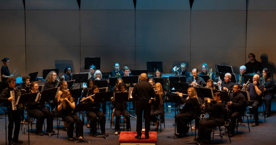  Members of the Rend Lake College concert band perform on stage during the 2026 Spring Concert: Chiaroscuro. Directed by conductor William Reynolds, who is shown from behind on a podium, the musicians play various instruments, including flutes, clarinets, and an accordion, in front of a neutral-toned backdrop