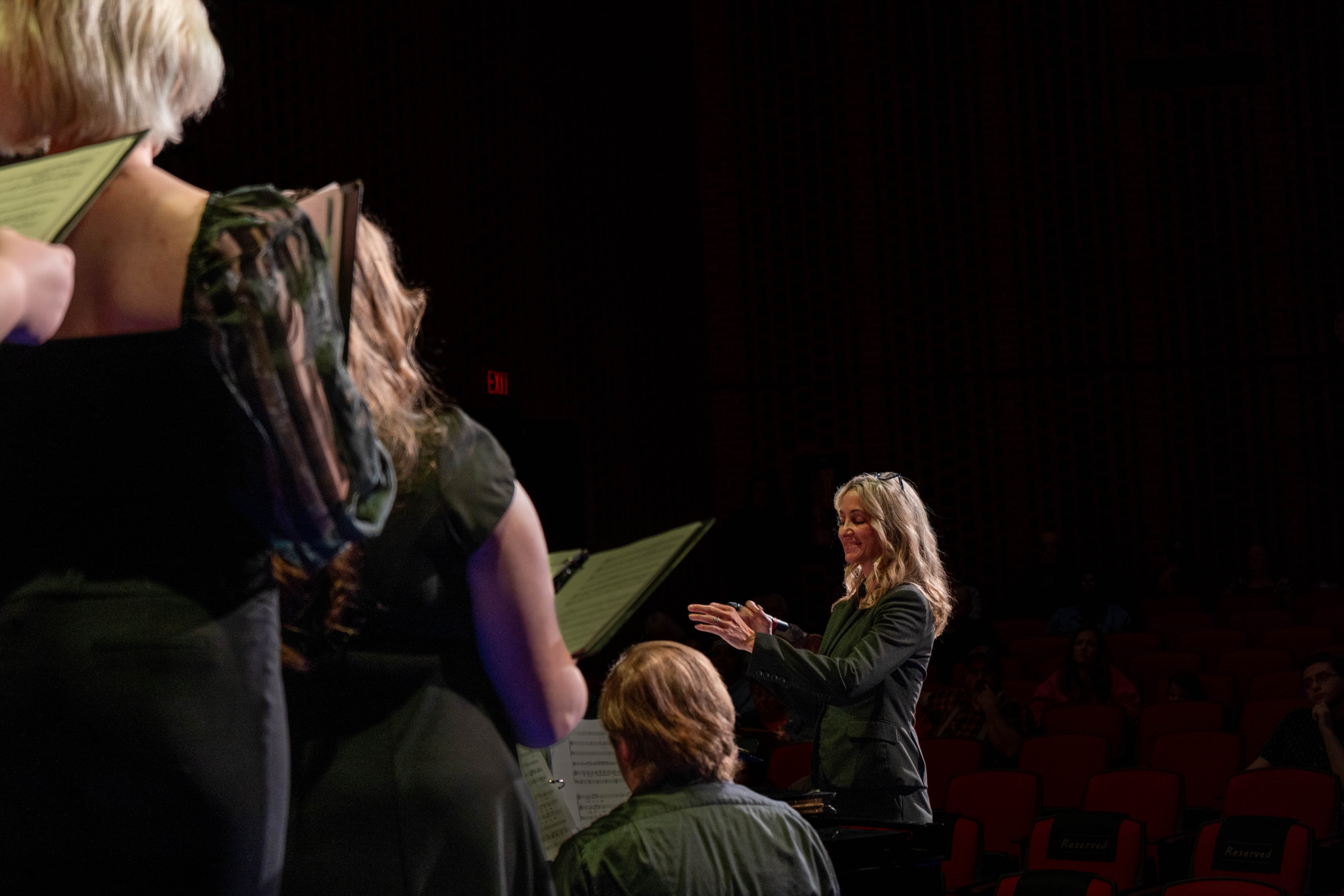 RLC music professor Sara Alstat-Sanders conducts the combined RLC and SIC choir during the 2026 Spring Concert: Chiaroscuro. The choir members stand in the foreground, facing the conductor as an audience watches from the darkened theatre seating.