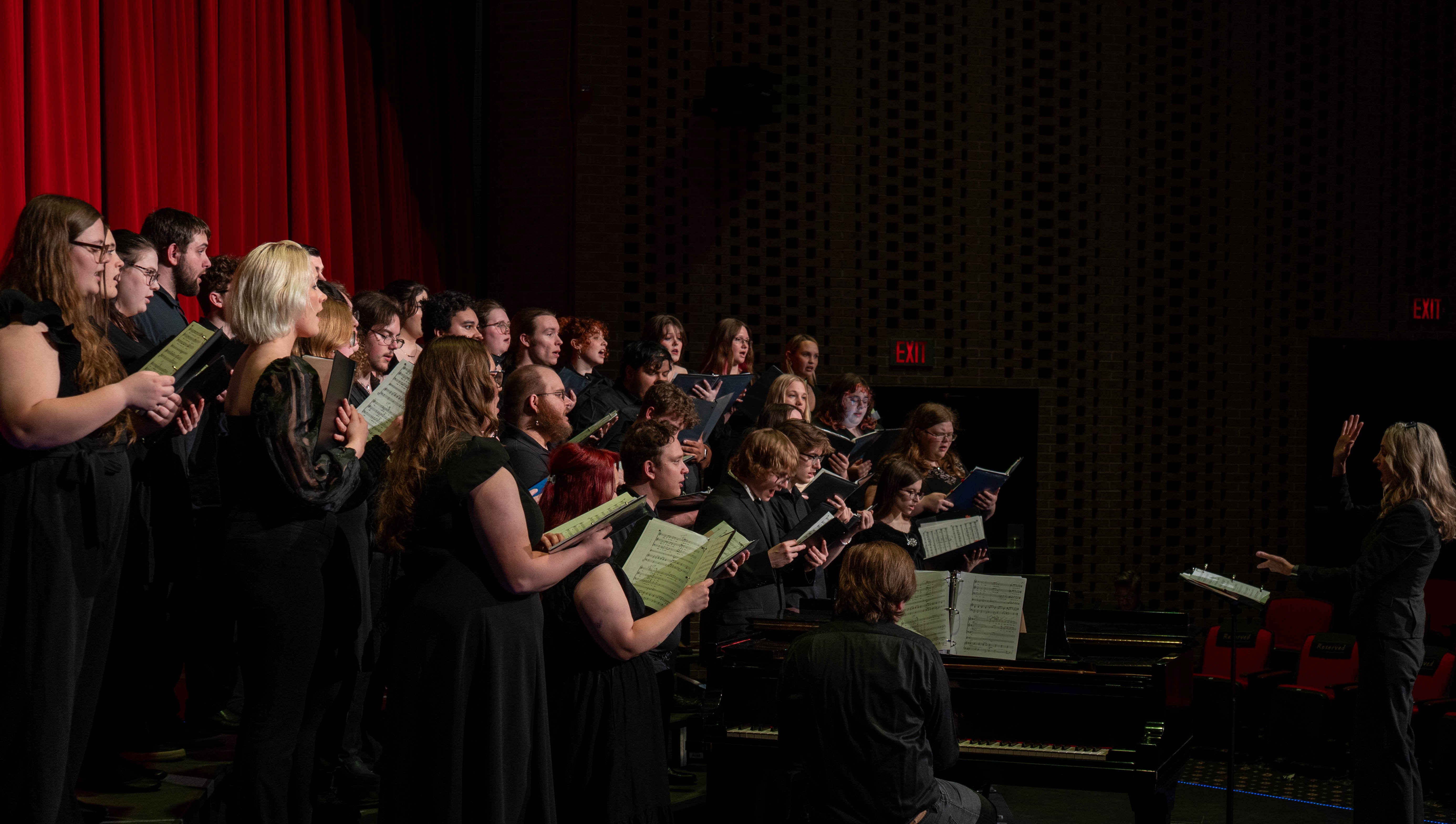 The combined RLC and SIC choir performs on stage during the 2026 Spring Concert: Chiaroscuro. Music professor Sara Alstat-Sanders conducts the large group of students, who are holding music folders and singing alongside a grand piano in the RLC Theatre.