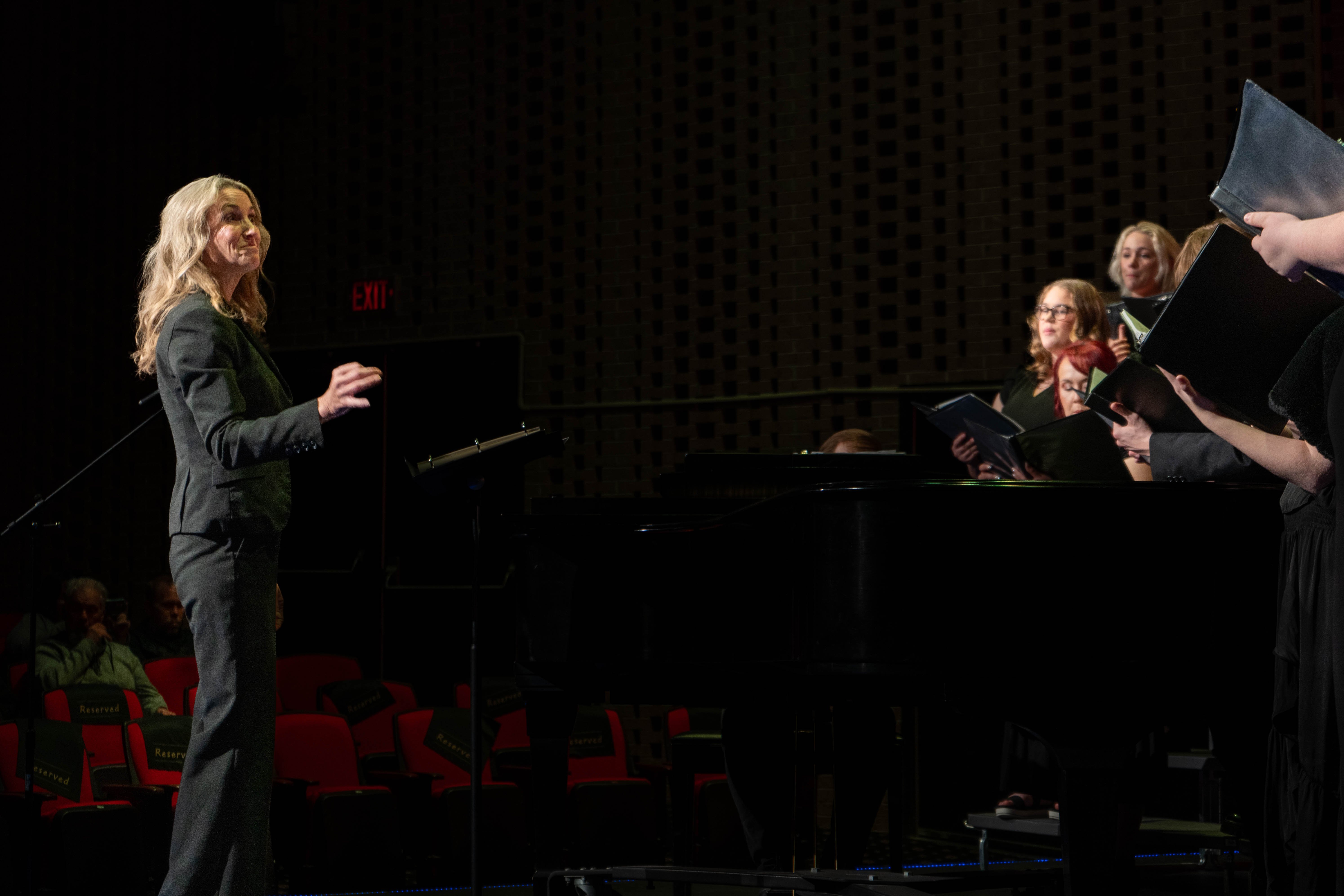 RLC music professor Sara Alstat-Sanders conducts the choir during the 2026 Spring Concert: Chiaroscuro. She stands on stage next to a grand piano, directing the students while an audience watches from the red seats of the RLC Theatre.