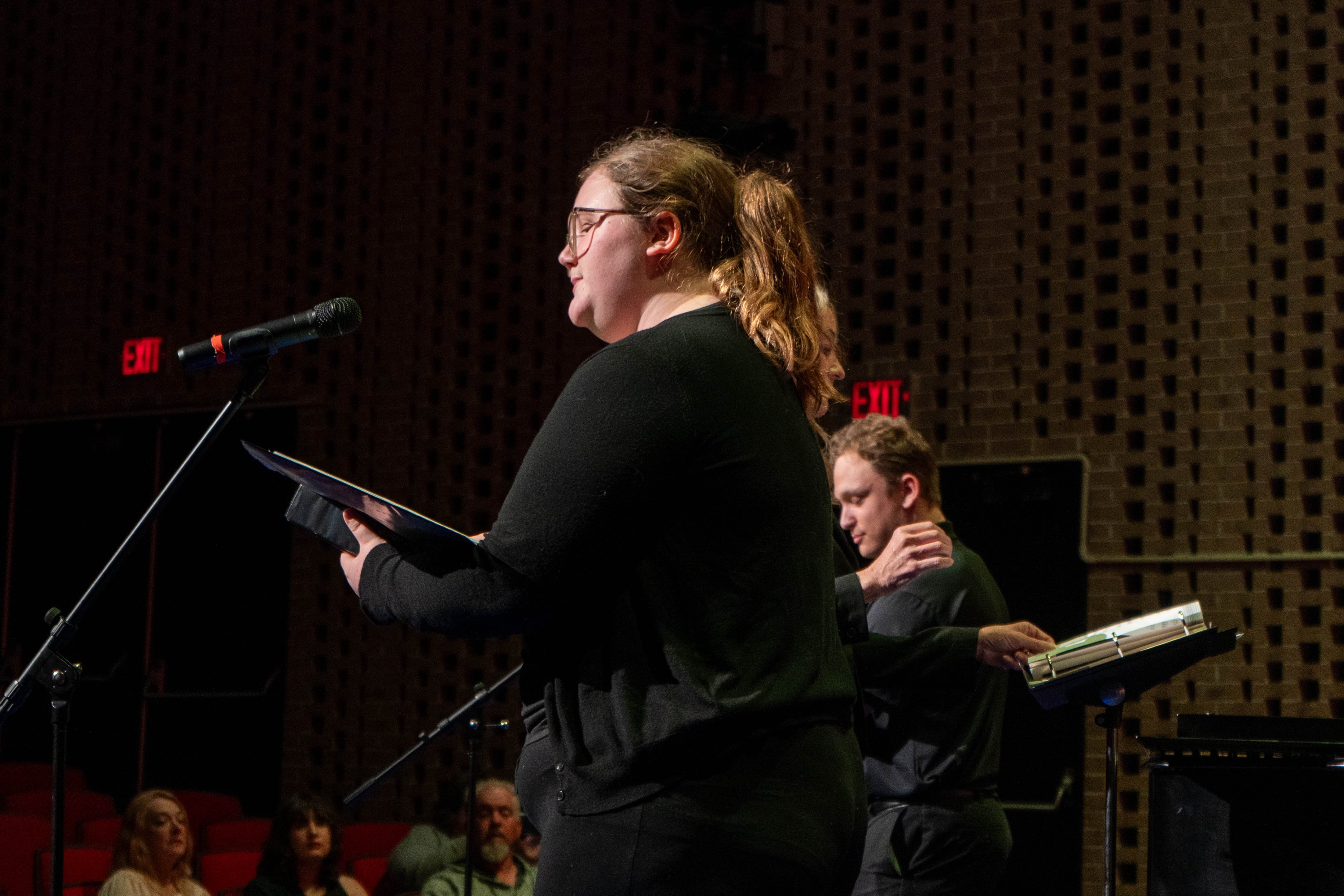 Victoria Bocock performs at a microphone during the 2026 Spring Concert: Chiaroscuro. She holds a music folder and sings on the RLC Theatre stage while other choir members and an audience are visible in the background.