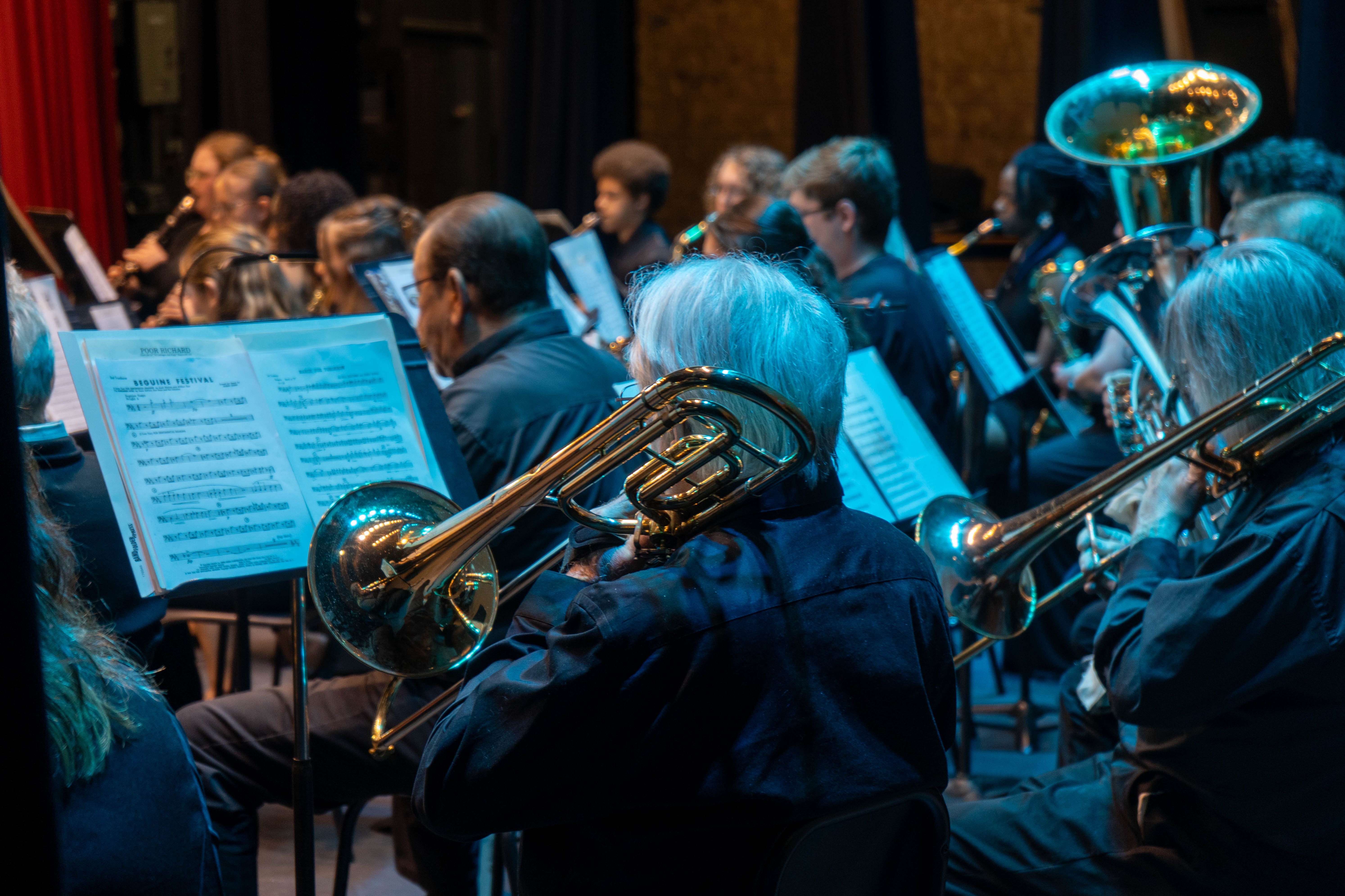 Trombone players and other members of the Rend Lake College concert band perform under blue stage lighting during the 2026 Spring Concert: Chiaroscuro. Sheet music for "Beguine Festival" is visible on a stand in the foreground.