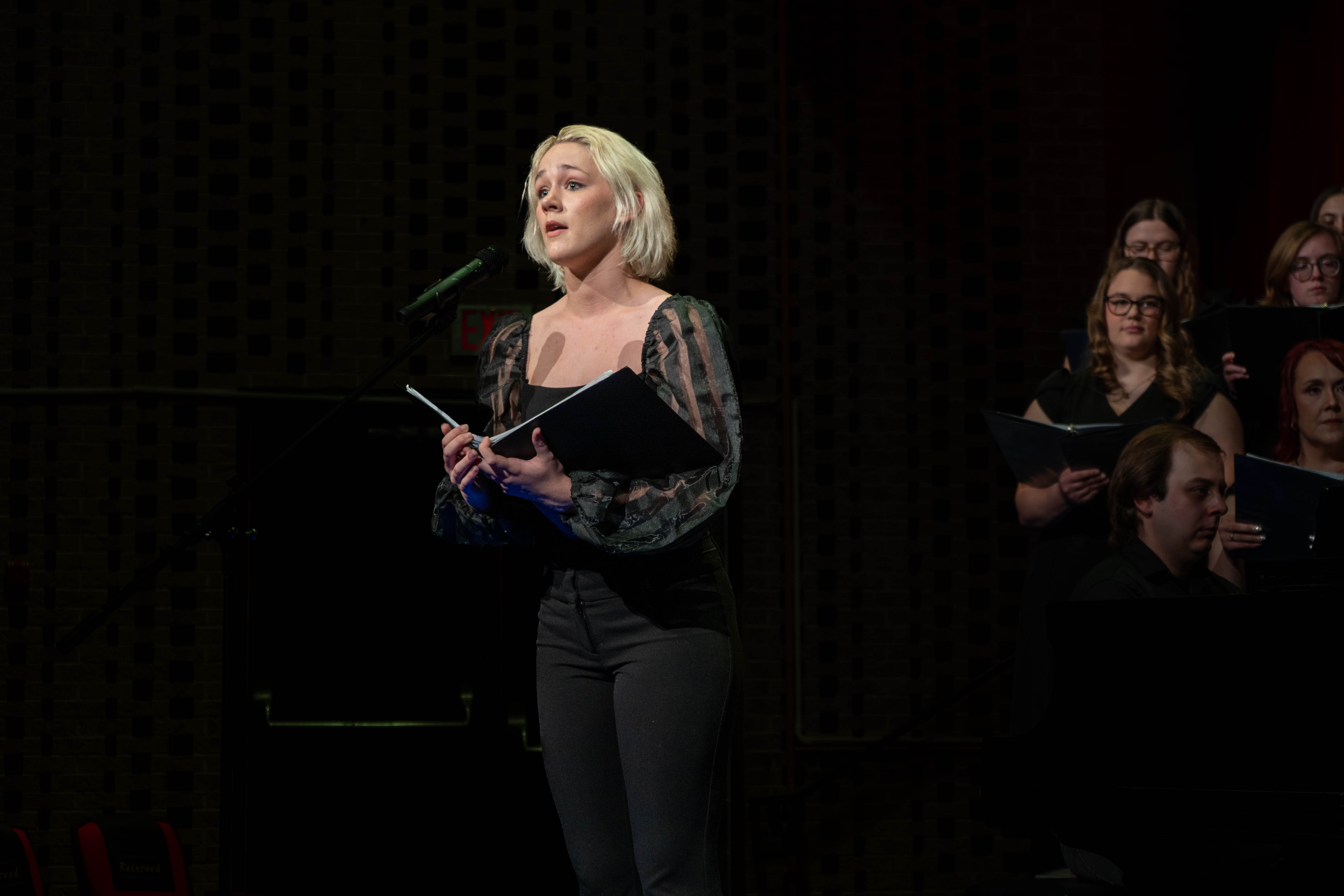Maddison Brower performs at a microphone during the 2026 Spring Concert: Chiaroscuro. She holds a music folder while members of the choir stand behind her on a dimly lit stage.
