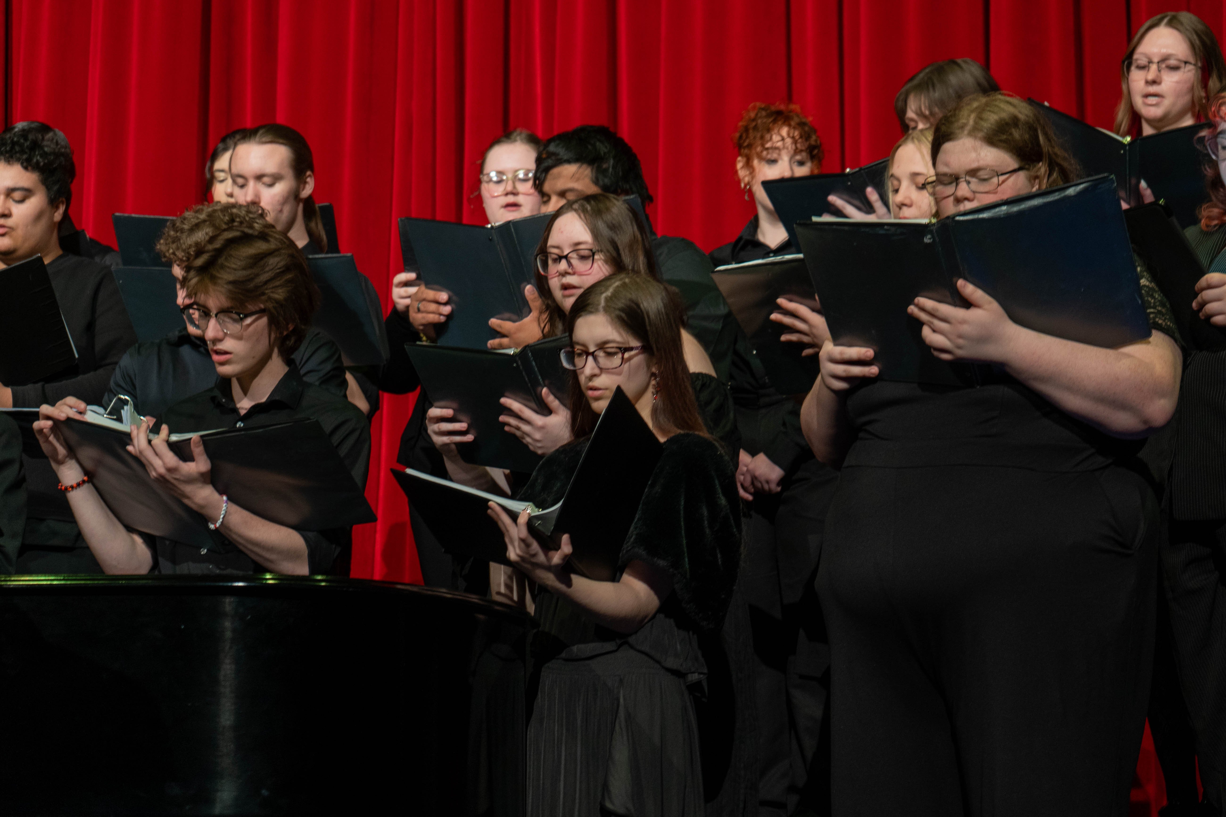 The combined RLC and SIC choir performs during the 2026 Spring Concert: Chiaroscuro. Dressed in black, the students hold music folders and sing in front of a vibrant red stage curtain at the RLC Theatre.
