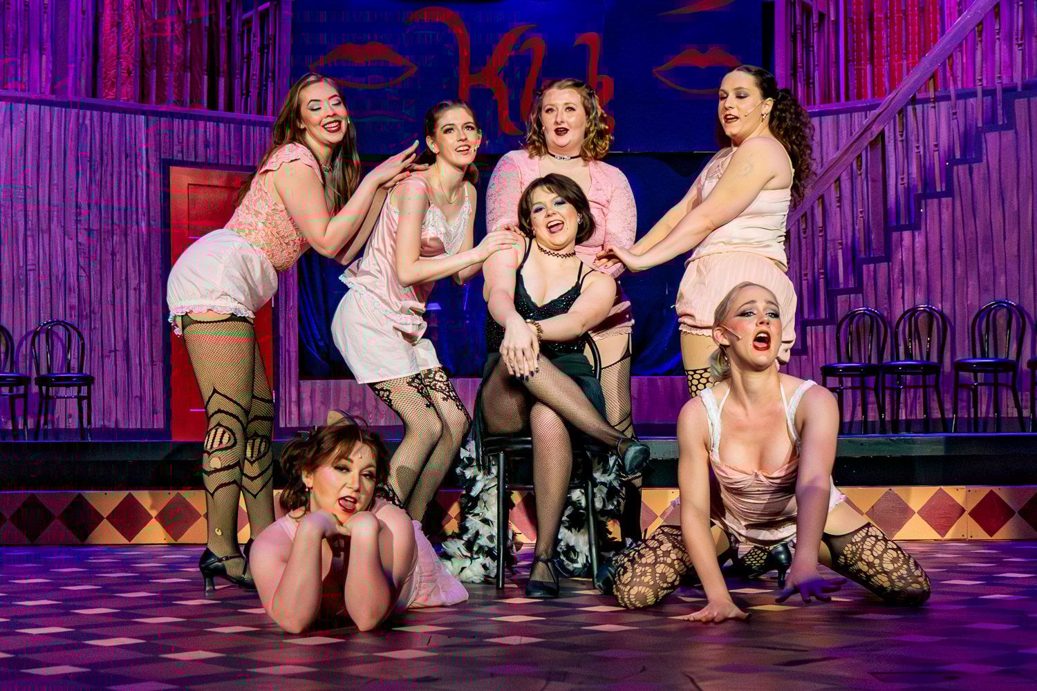 Cadence Morgan as Sally Bowles sits center stage on a black chair, surrounded by six Kit Kat Girls in a posed ensemble.