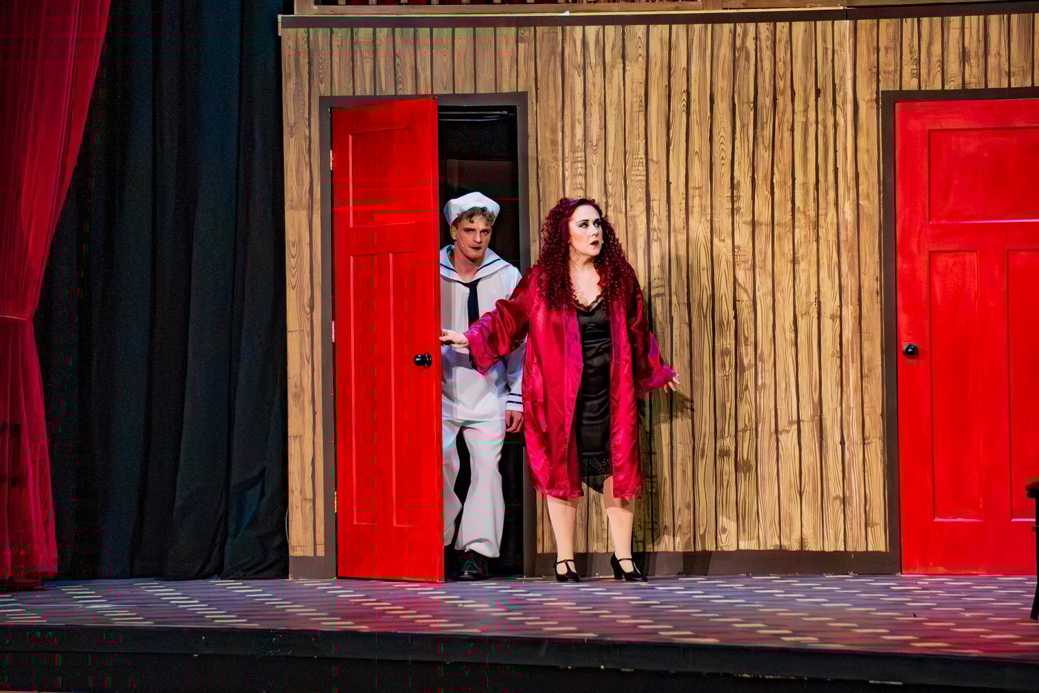 Fräulein Kost stands on stage in a black slip and a vibrant red silk robe, looking toward the audience with a dramatic expression. Behind her, a sailor in a white uniform and hat peers out from a bright red doorway in a wooden-paneled set