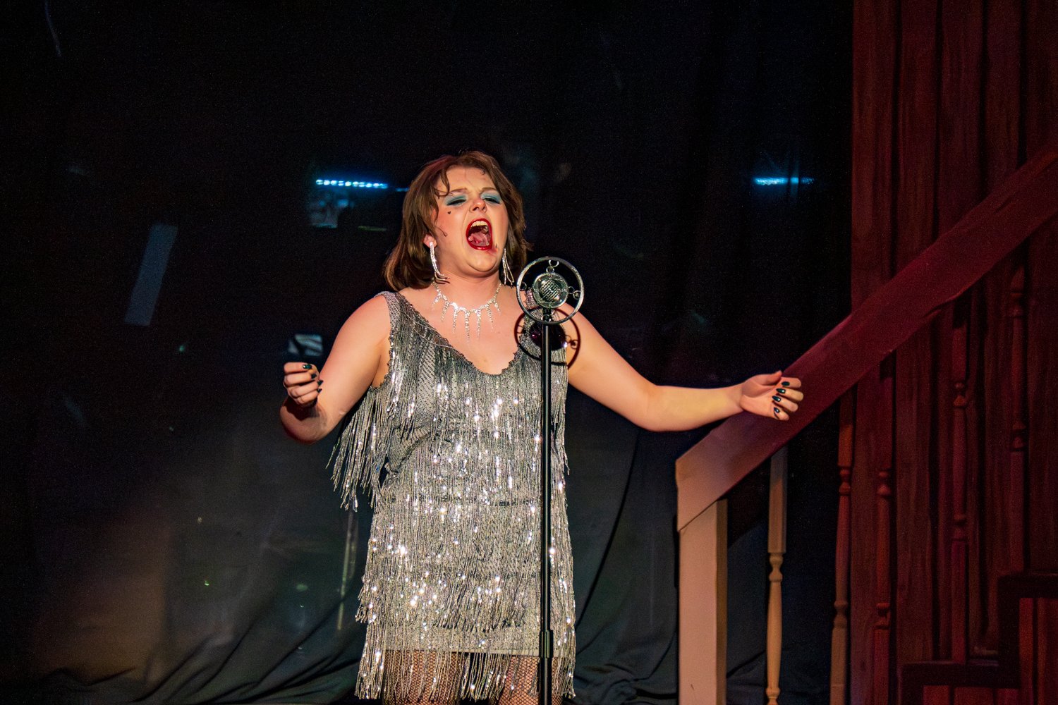 Cadence Morgan as Sally Bowles delivers a powerful, emotional performance of the title song, "Cabaret." She is dressed in a shimmering silver-fringed flapper dress, accessorized with a sparkling necklace and long earrings.