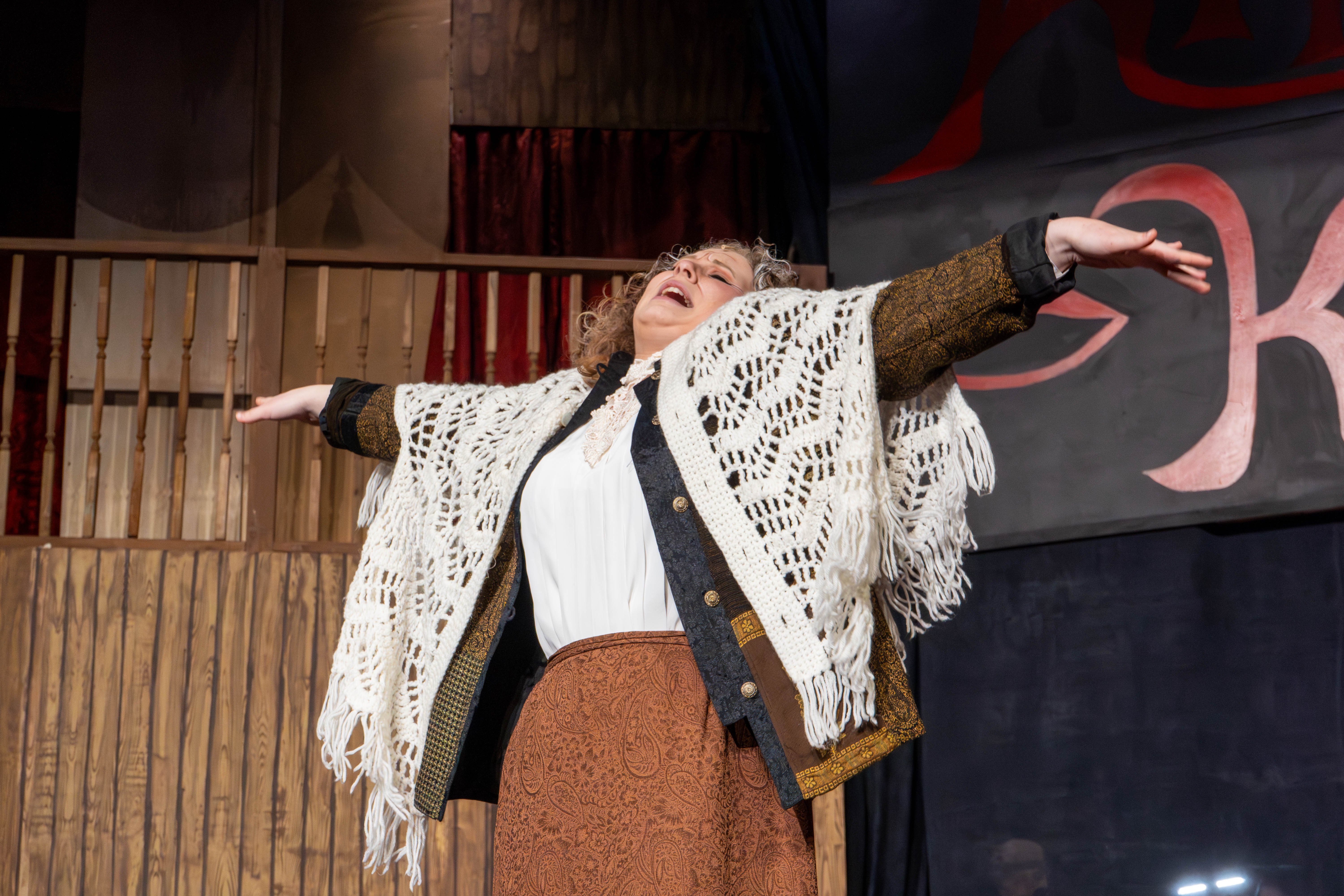 Actor Jinx Craddock portraying Fräulein Schneider from the musical Cabaret stands on stage with her arms outstretched and eyes closed in an emotional, expressive pose.