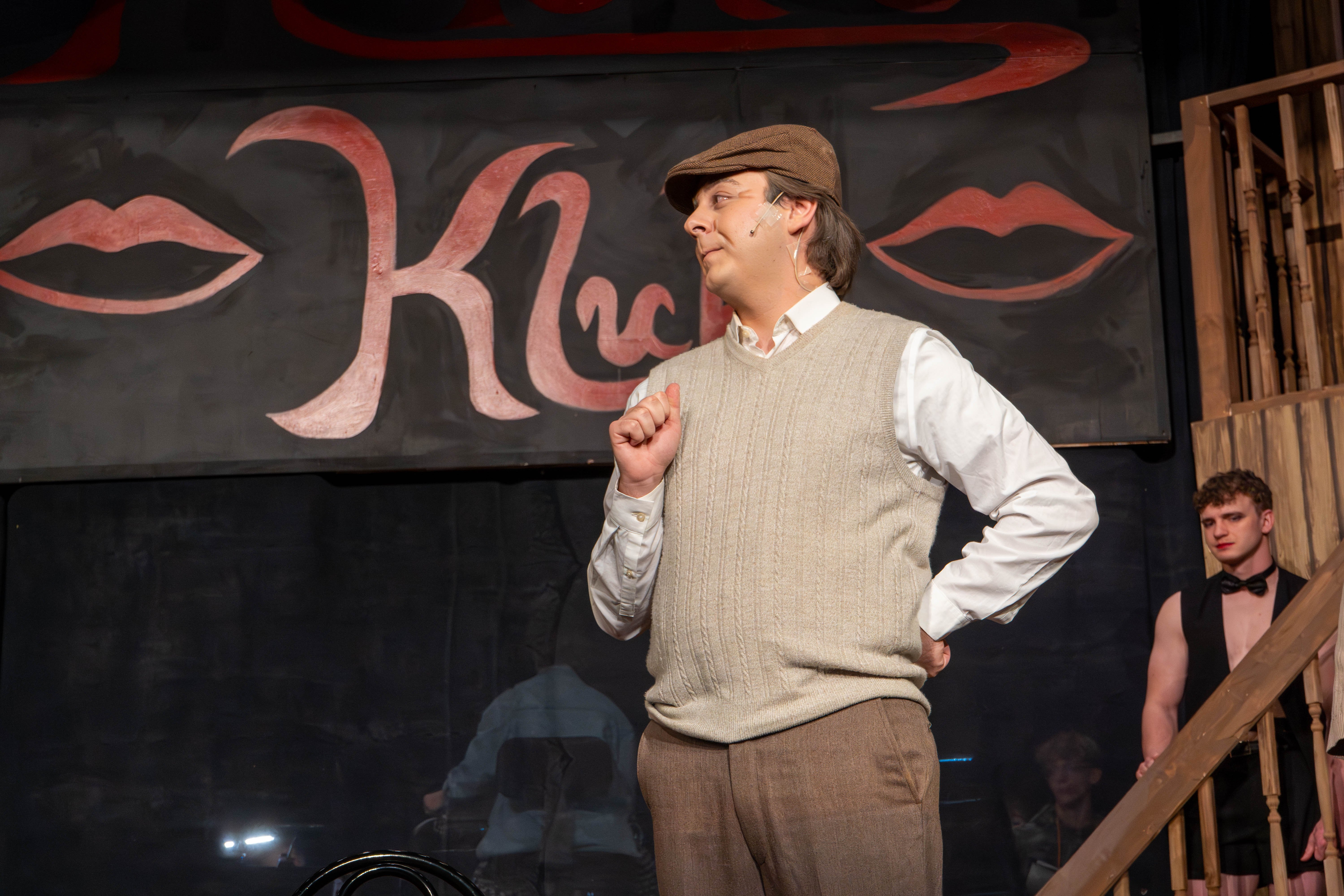 Herr Schultz stands on stage wearing a brown flat cap, a white collared shirt, a tan sweater vest, and brown trousers