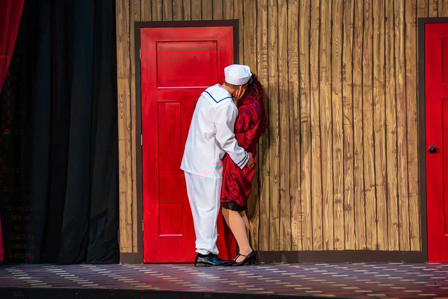 An actor portraying a sailor in a white uniform and a performer as Fräulein Kost in a red silk robe share a dramatic kiss on stage.