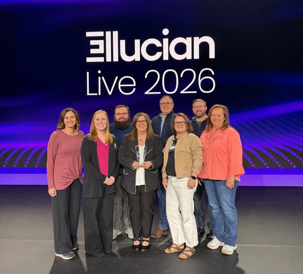  A group of eight people from the Rend Lake College Ellucian team posing on a stage at the Ellucian Live 2026 conference. They are standing in front of a large digital screen displaying the "Ellucian Live 2026" logo on a dark purple background. In the center, RLC President Dr. Lori Ragland holds a clear glass trophy.  The group includes:  Front row (L-R): Dean of Institutional Research Amy Epplin, Bursar & Director of Accounting Mallory Howell, RLC President Dr. Lori Ragland, Dean of Enrollment Services Jena Jensik, and Payroll & Benefits Accountant Glenna Maxwell.  Back row (L-R): Software Developer/System Admin Andrew Lingle, Vice President of Academic & Student Services Buster Leeck, and Software Developer/System Admin Cory Braden.