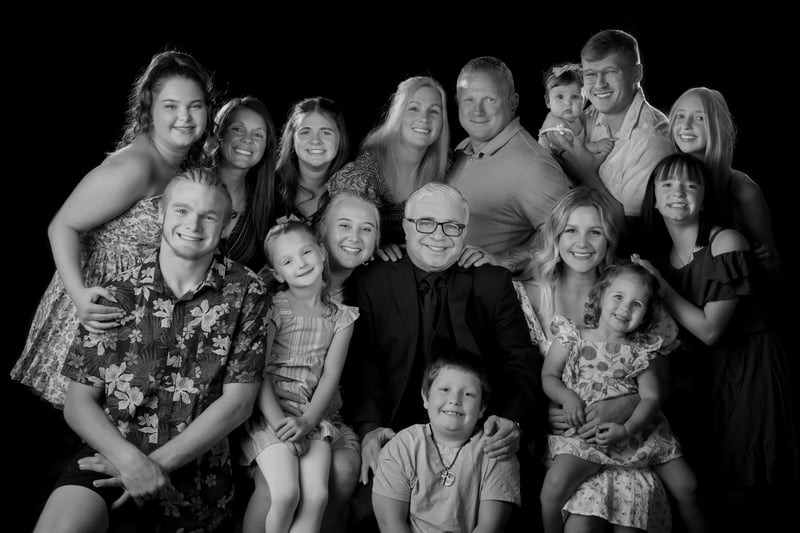 A black-and-white studio portrait of a large multi-generational family, including adults, teenagers, children, and an infant, posing together against a dark background.