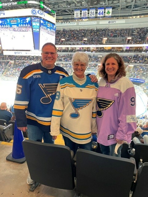 hree people standing together in the stands of an indoor hockey arena, each wearing a different St. Louis Blues hockey jersey and smiling at the camera.